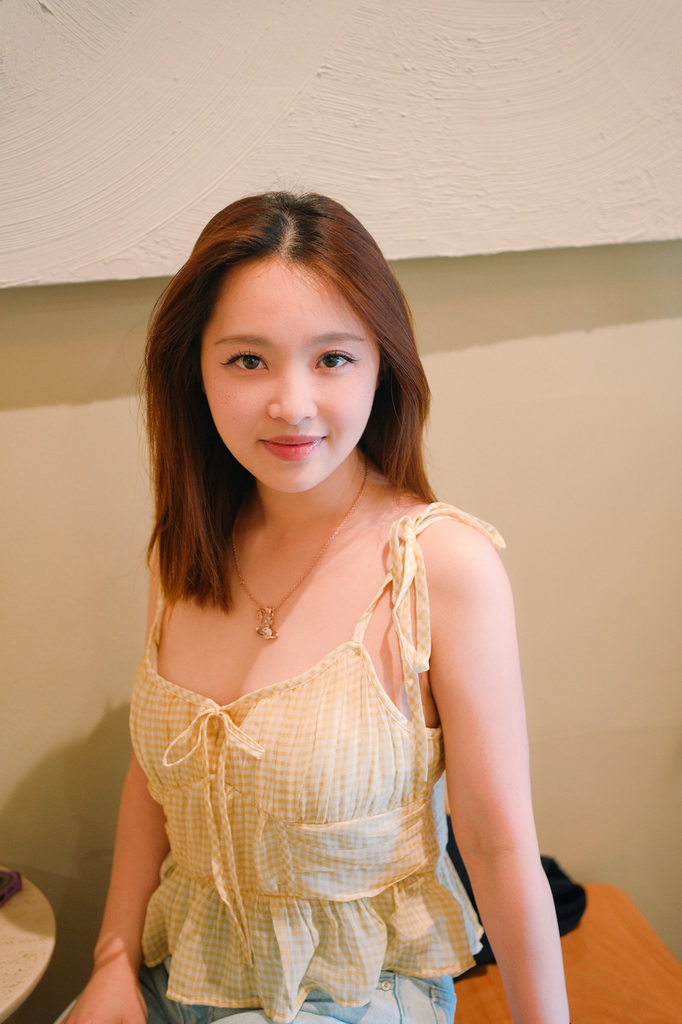 A young woman with shoulder-length brown hair, wearing a yellow checkered spaghetti strap top, smiling at the camera. She is standing in a room with beige walls, with a table and some objects partially visible in the background.