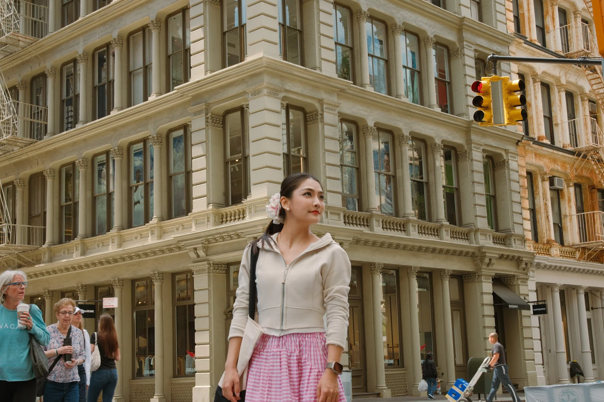 City street scene with a woman in a pink and white checkered skirt and beige jacket, several pedestrians, and a historic building with rounded windows and ornate architectural details under a traffic light showing red.