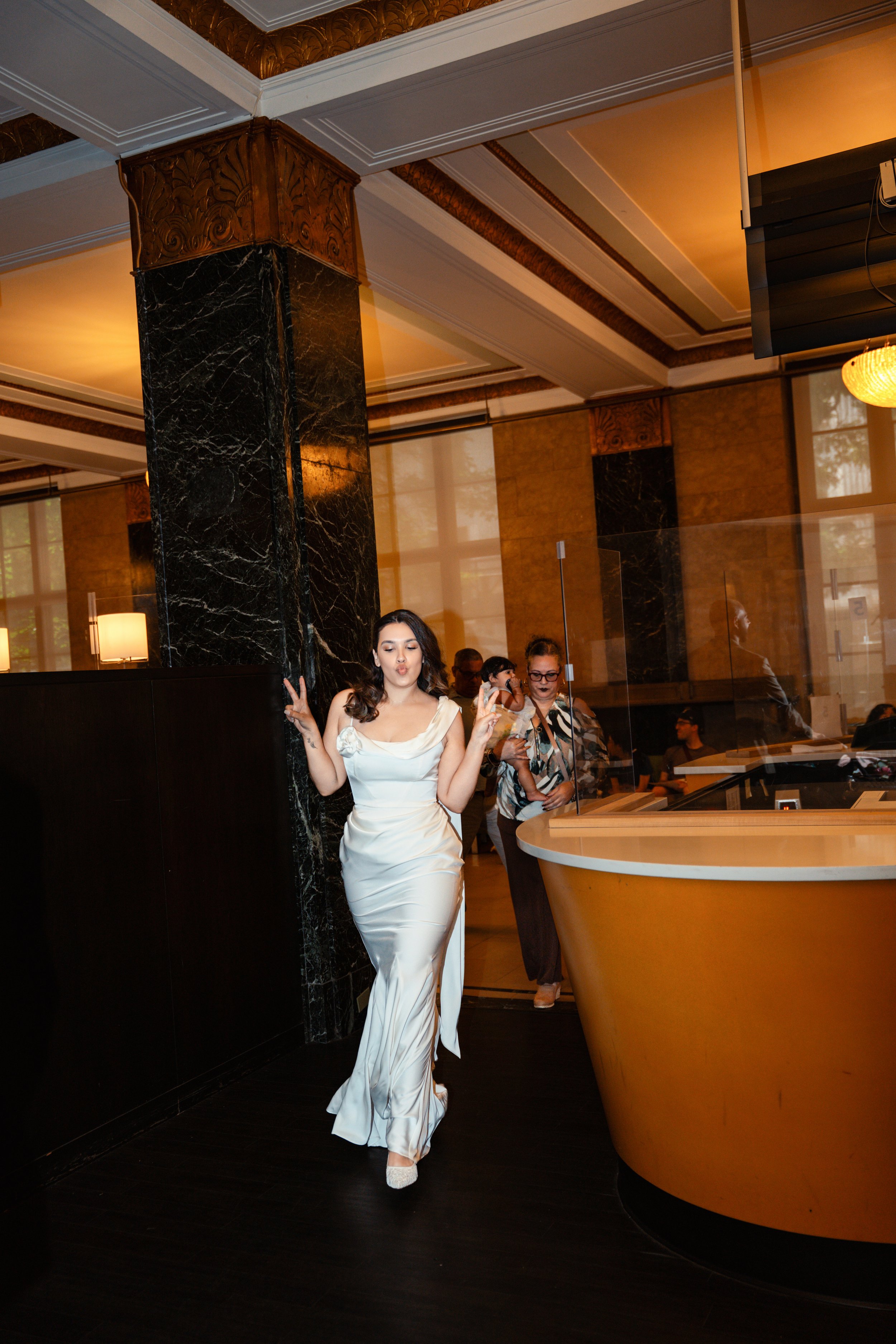 A woman in an elegant white satin gown making a peace sign and puckering her lips while standing in a fancy restaurant or hotel lobby. Multiple people are visible in the background, some standing and some seated. The interior features a large black m