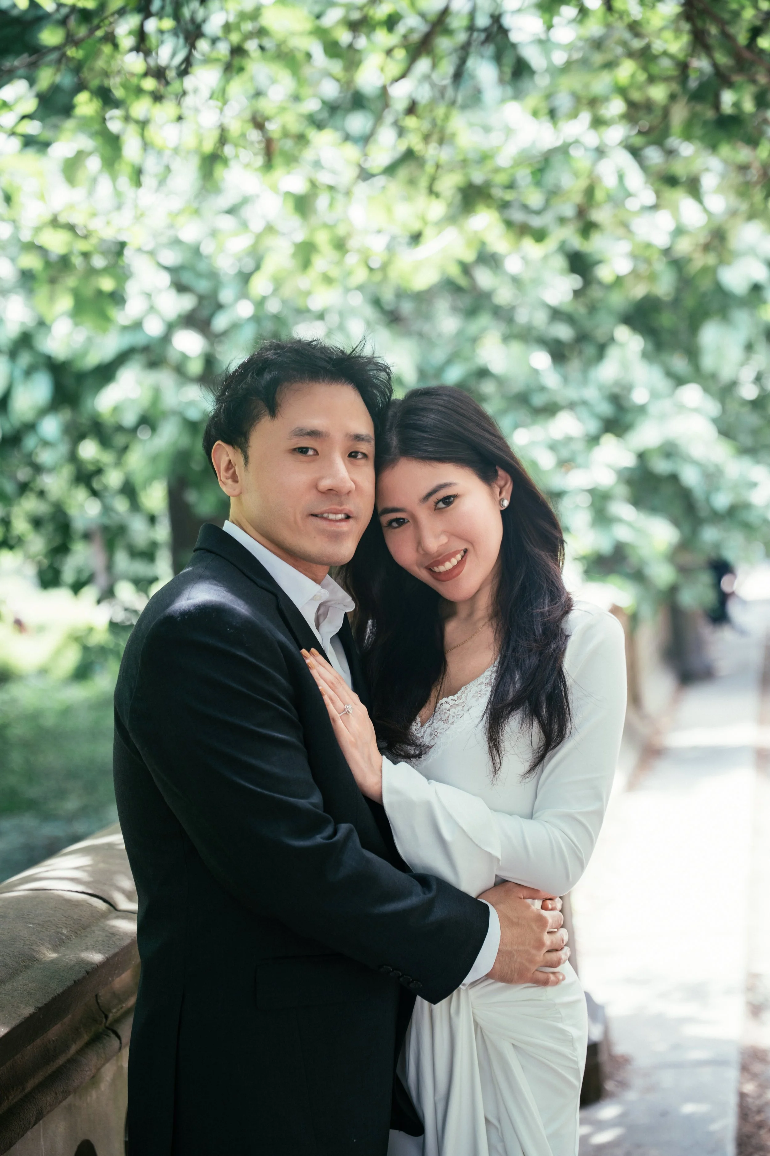 A couple dressed in formal attire, standing close together outdoors with green foliage in the background.