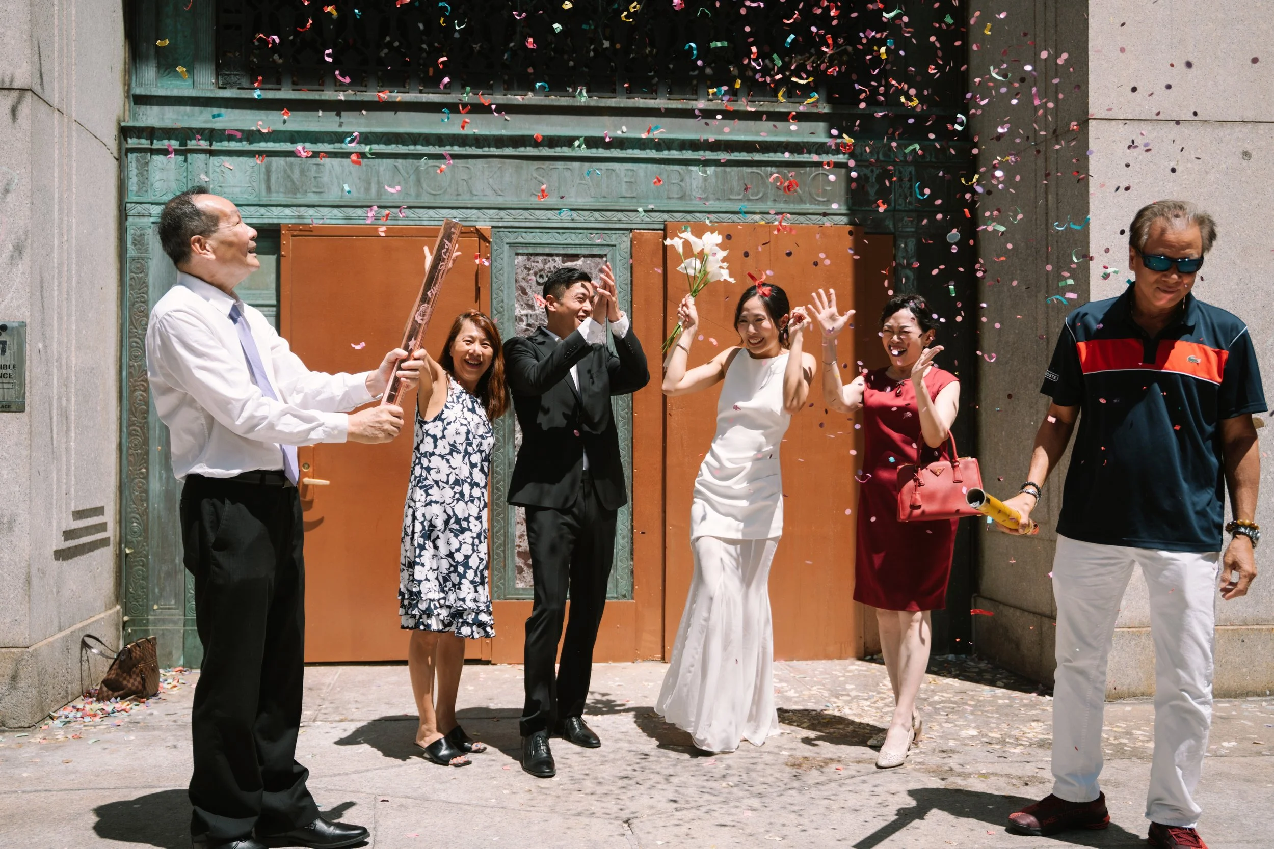 Group of five people celebrating outside, colorful confetti falling, some holding flowers, smiling and having fun.
