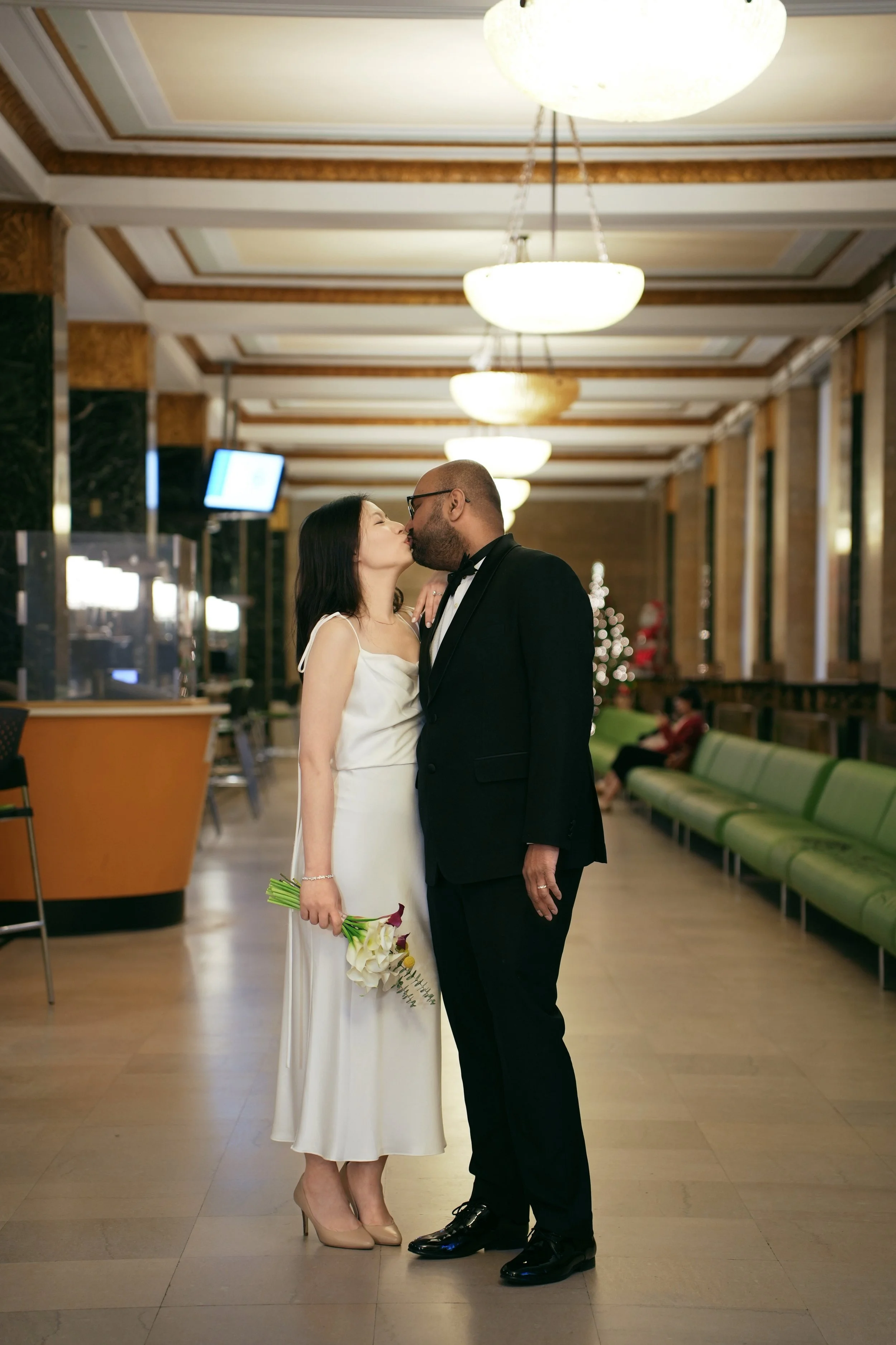 A newlywed couple is sharing a kiss in a spacious, well-lit hotel lobby. The bride wears a white dress and holds a bouquet of flowers, while the groom is in a black tuxedo. The background features a Christmas tree, green benches, and decorative light