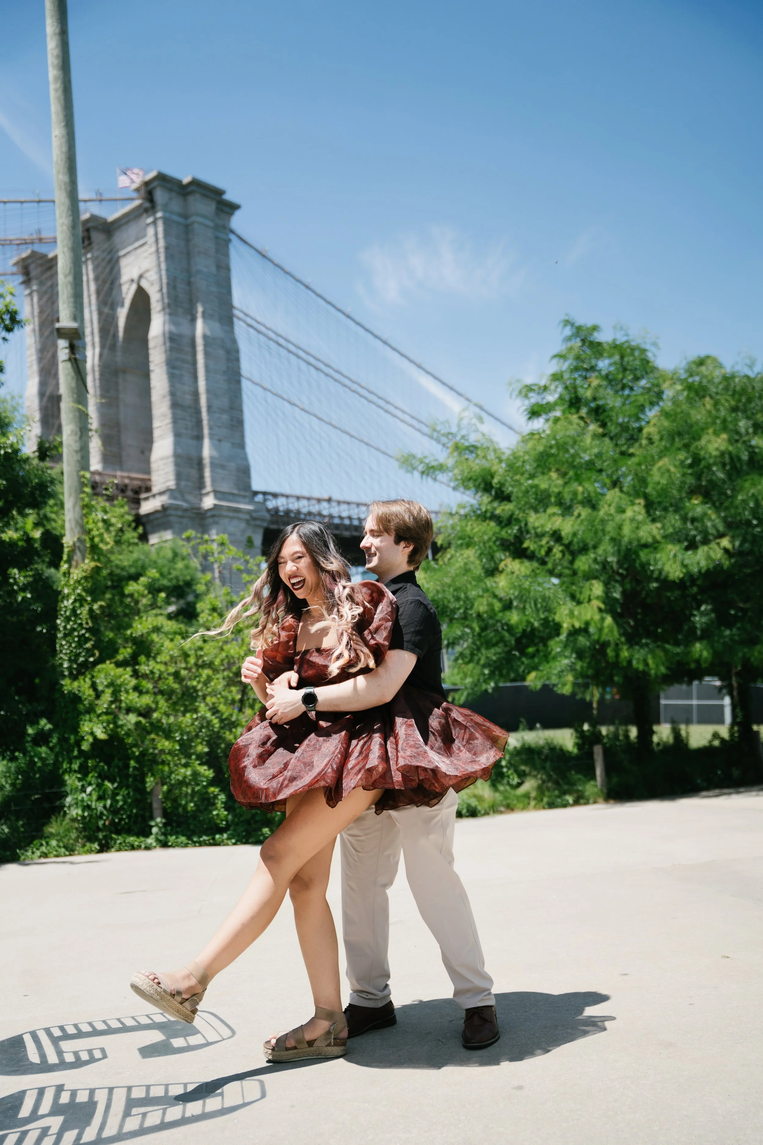 A man in beige pants and black shoes holding a woman in a brown dress with a floral pattern, both laughing and dancing on a sidewalk, with the Brooklyn Bridge, green trees, and a clear blue sky in the background.