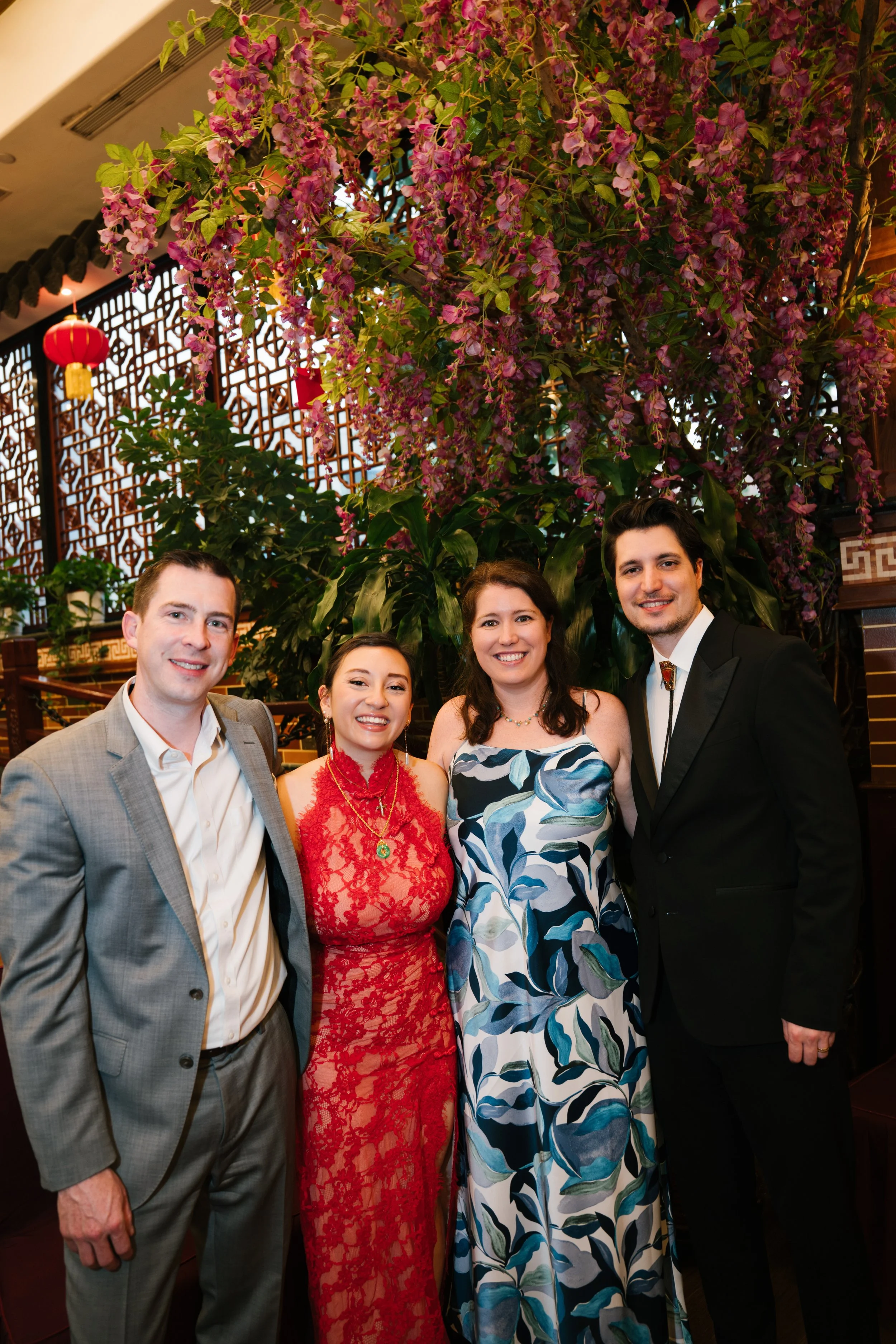 Five people are standing together indoors, smiling. They are dressed in formal and semi-formal attire with a large flowering plant and decorative wood panels in the background.