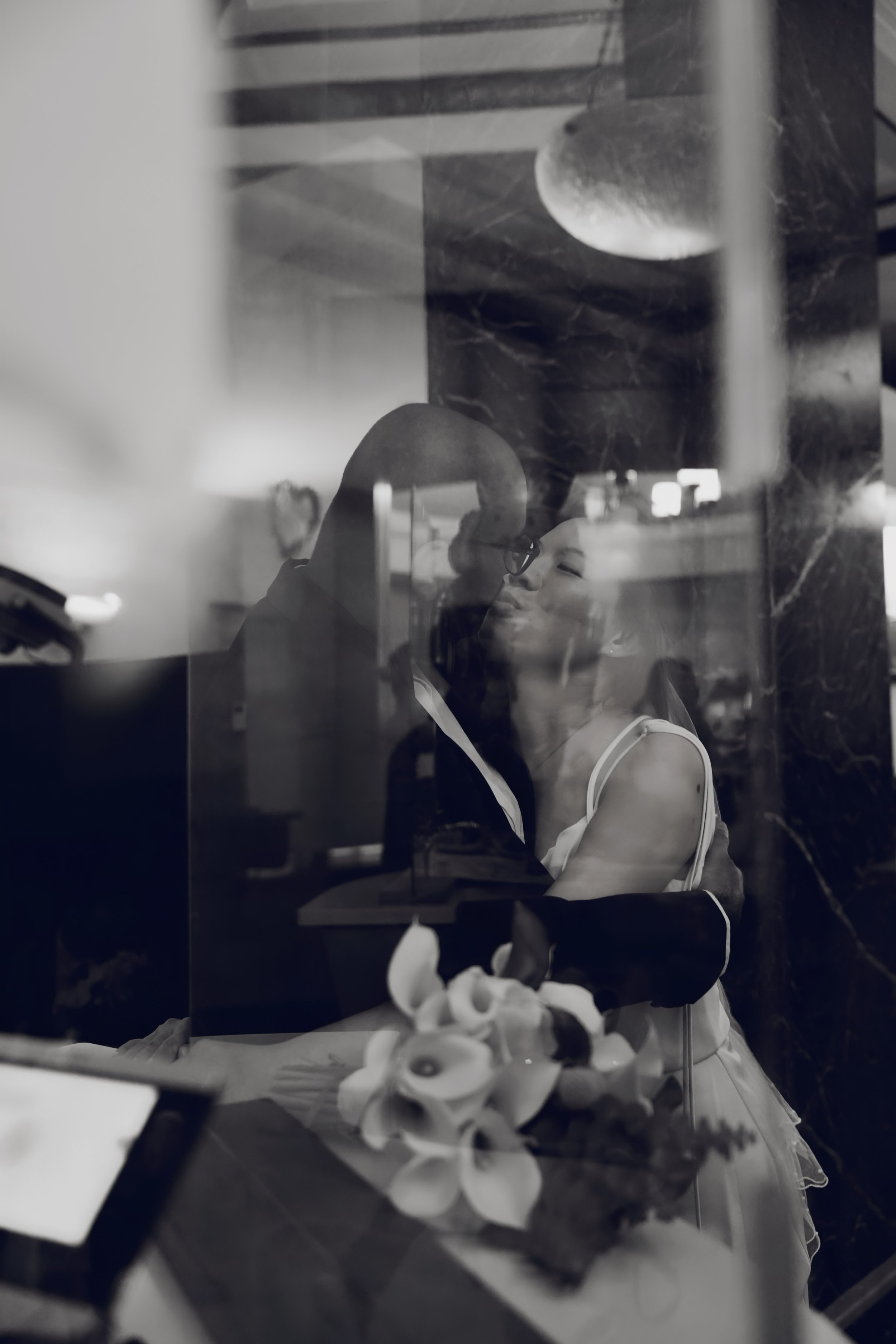 Reflection of a man sitting at a table with a flower arrangement, as seen through a glass window.