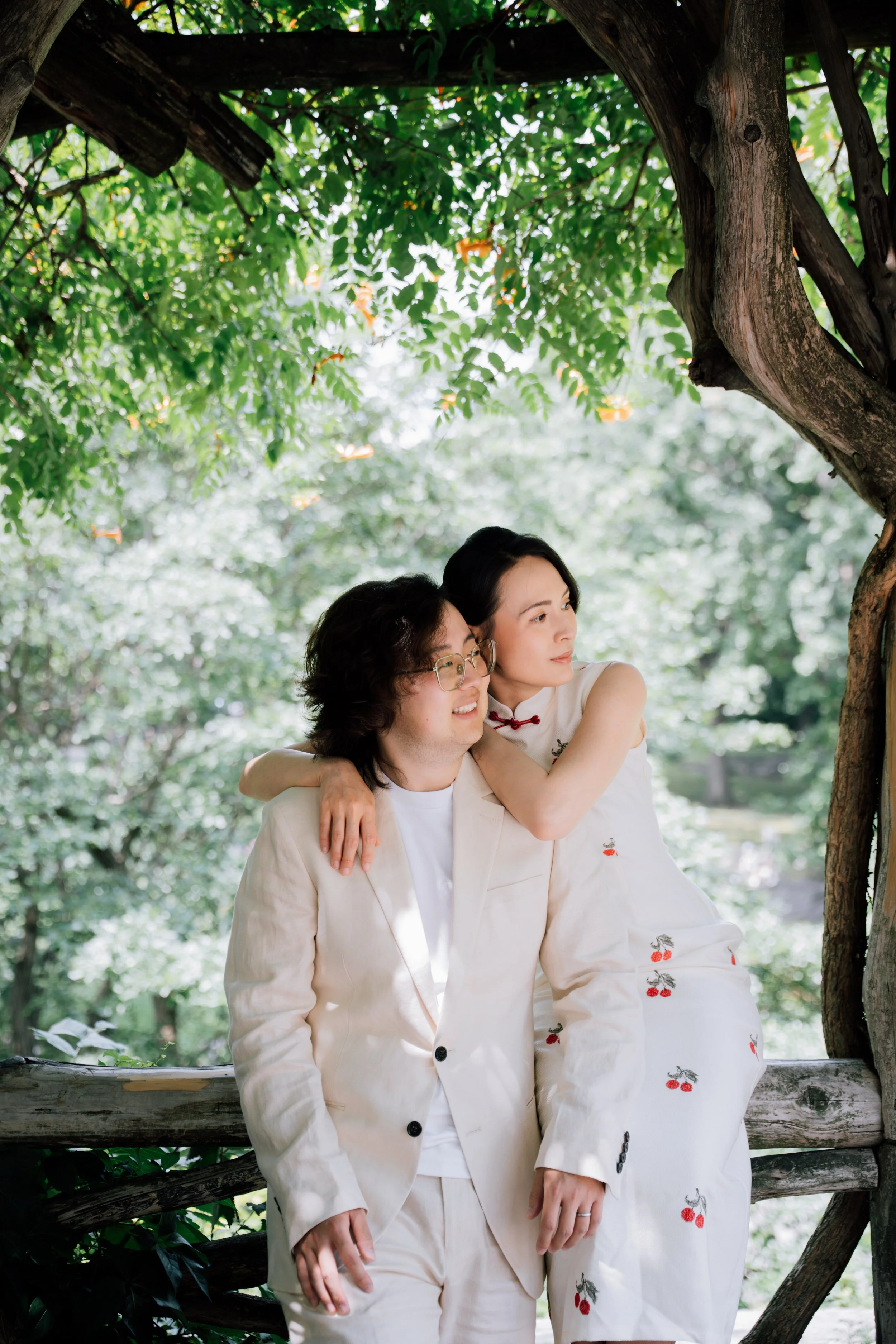 Two people, a man and a woman, embracing and smiling under a tree in a lush outdoor setting.
