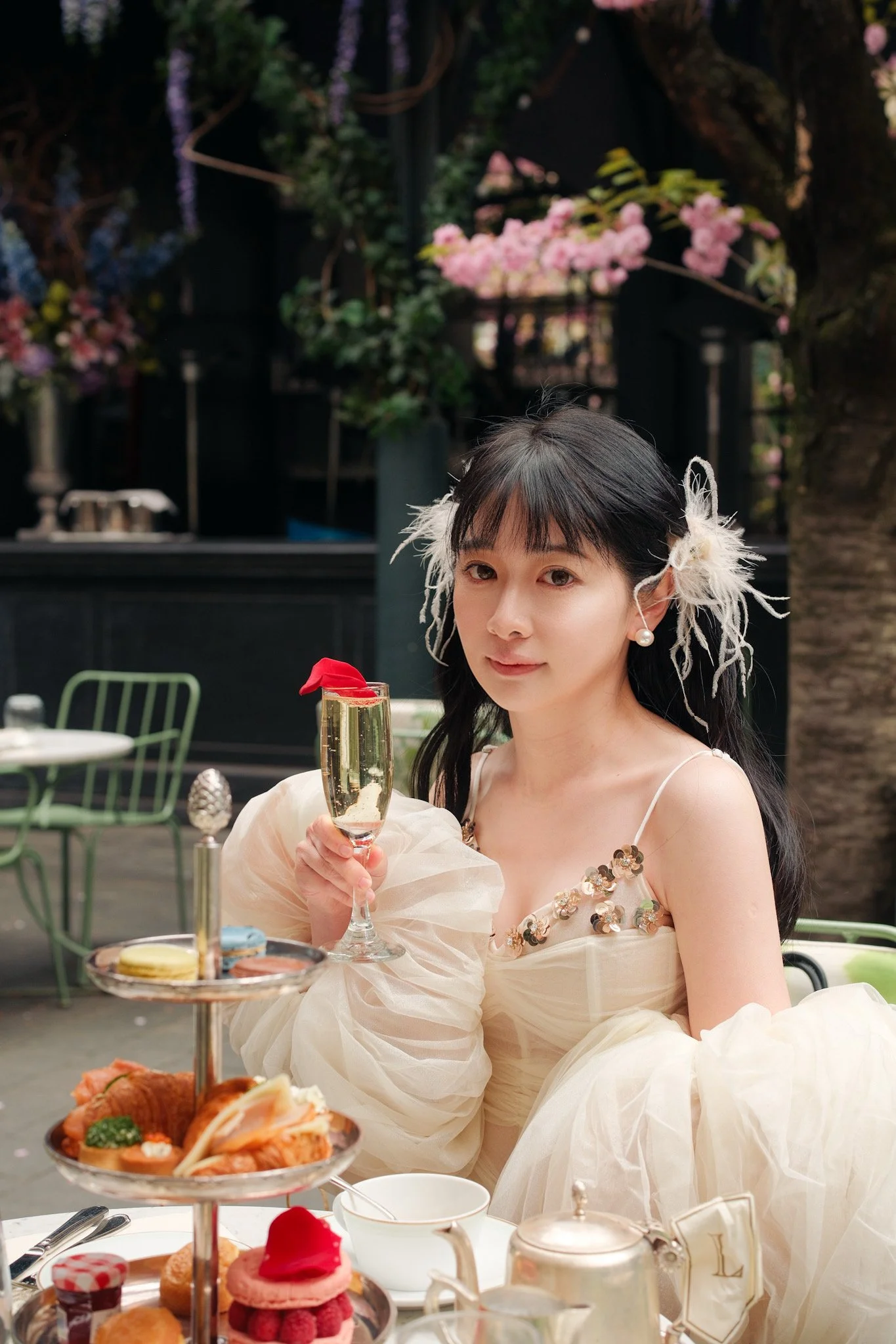 A woman dressed in an elegant cream-colored gown with sequin embellishments and feathered accessories in her hair, holding a glass of champagne at a high tea or afternoon tea setting, with a tiered tray of desserts in the foreground and floral decora