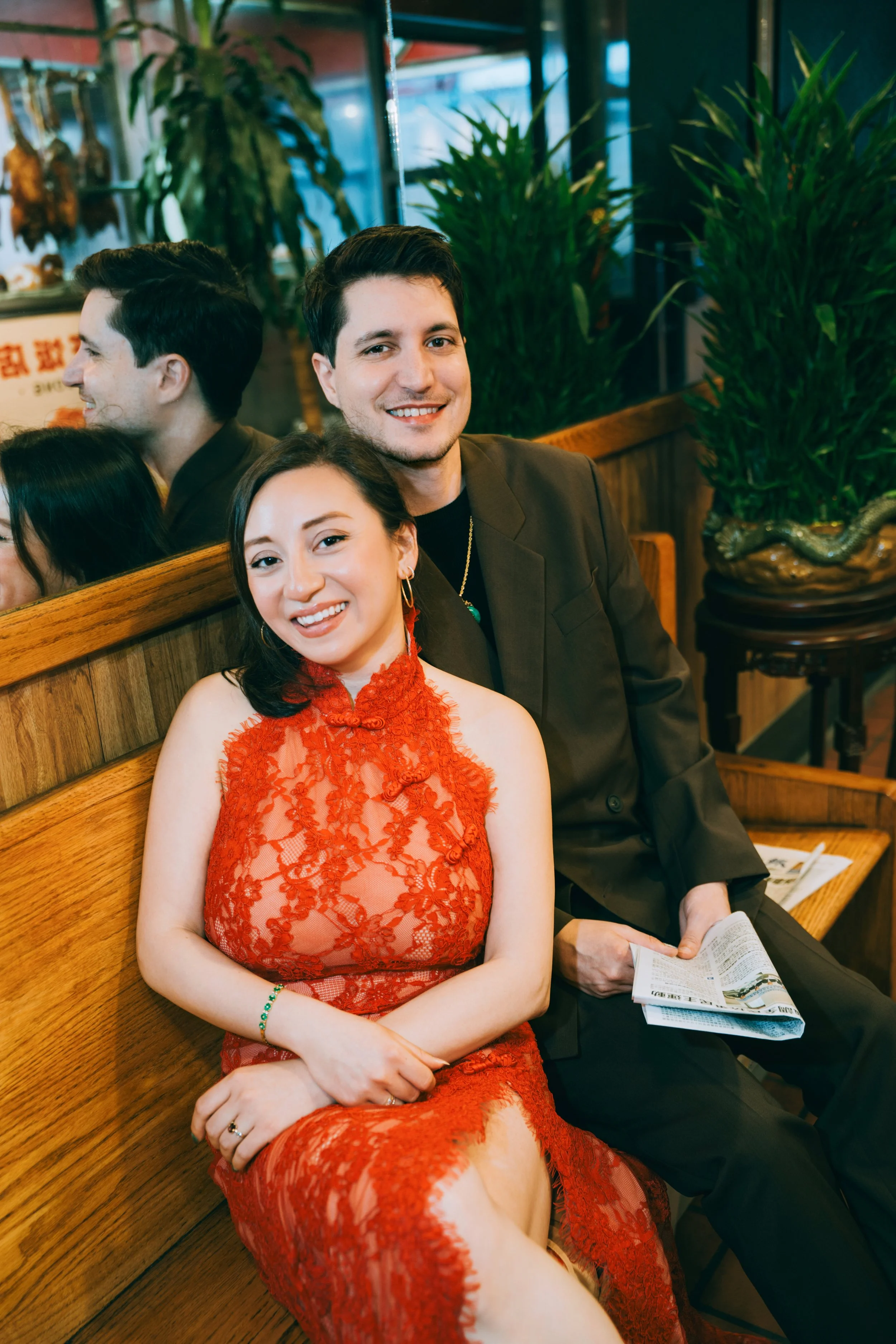 A young man and woman sitting on a wooden bench inside a restaurant, smiling at the camera. The woman wears a red lace dress, and the man is dressed in a dark blazer. They are surrounded by large green potted plants and a mirror reflecting part of th