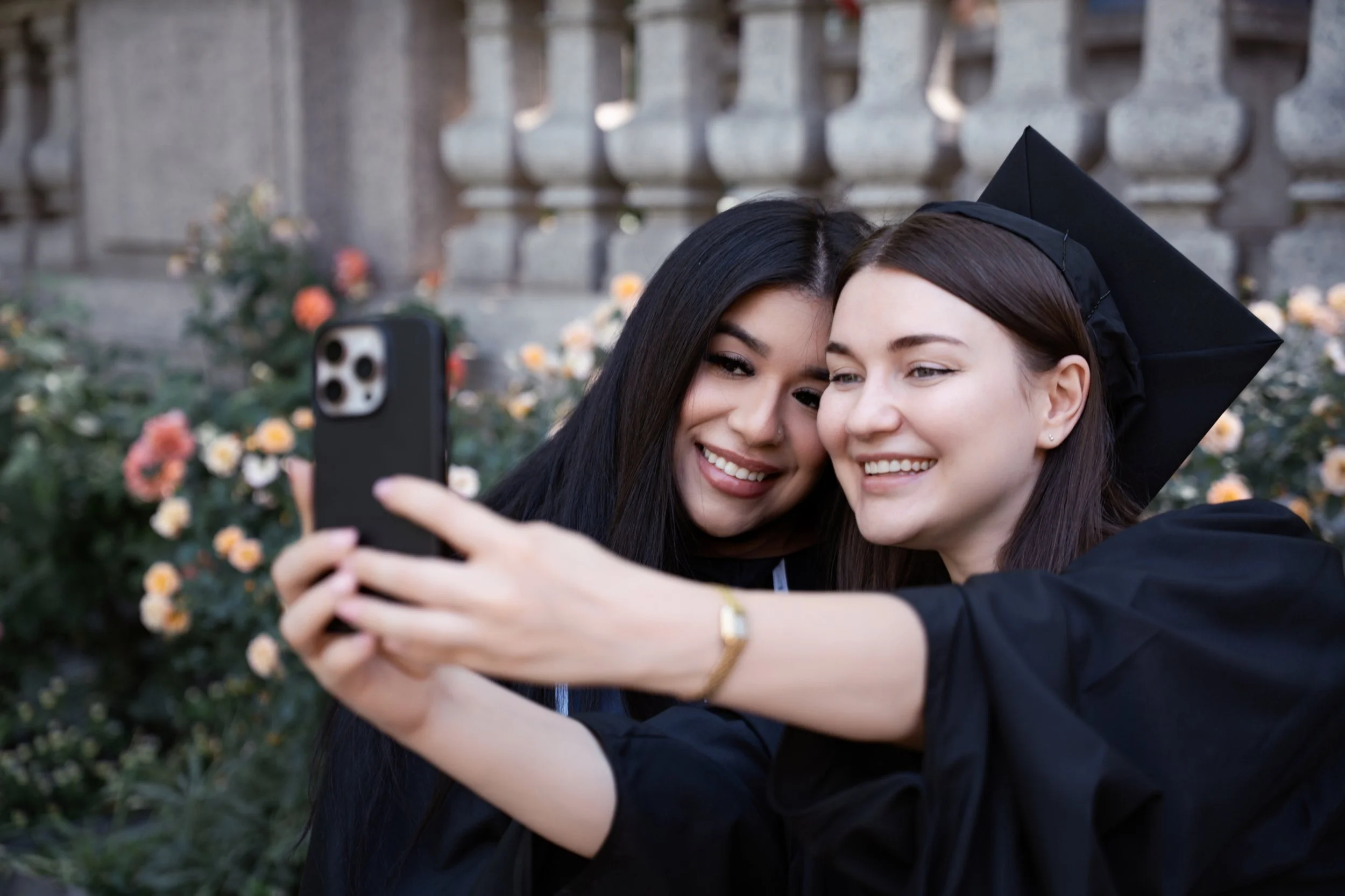 Two young women in graduation cap and gown taking a selfie outside, with flowers and a stone fence in the background.