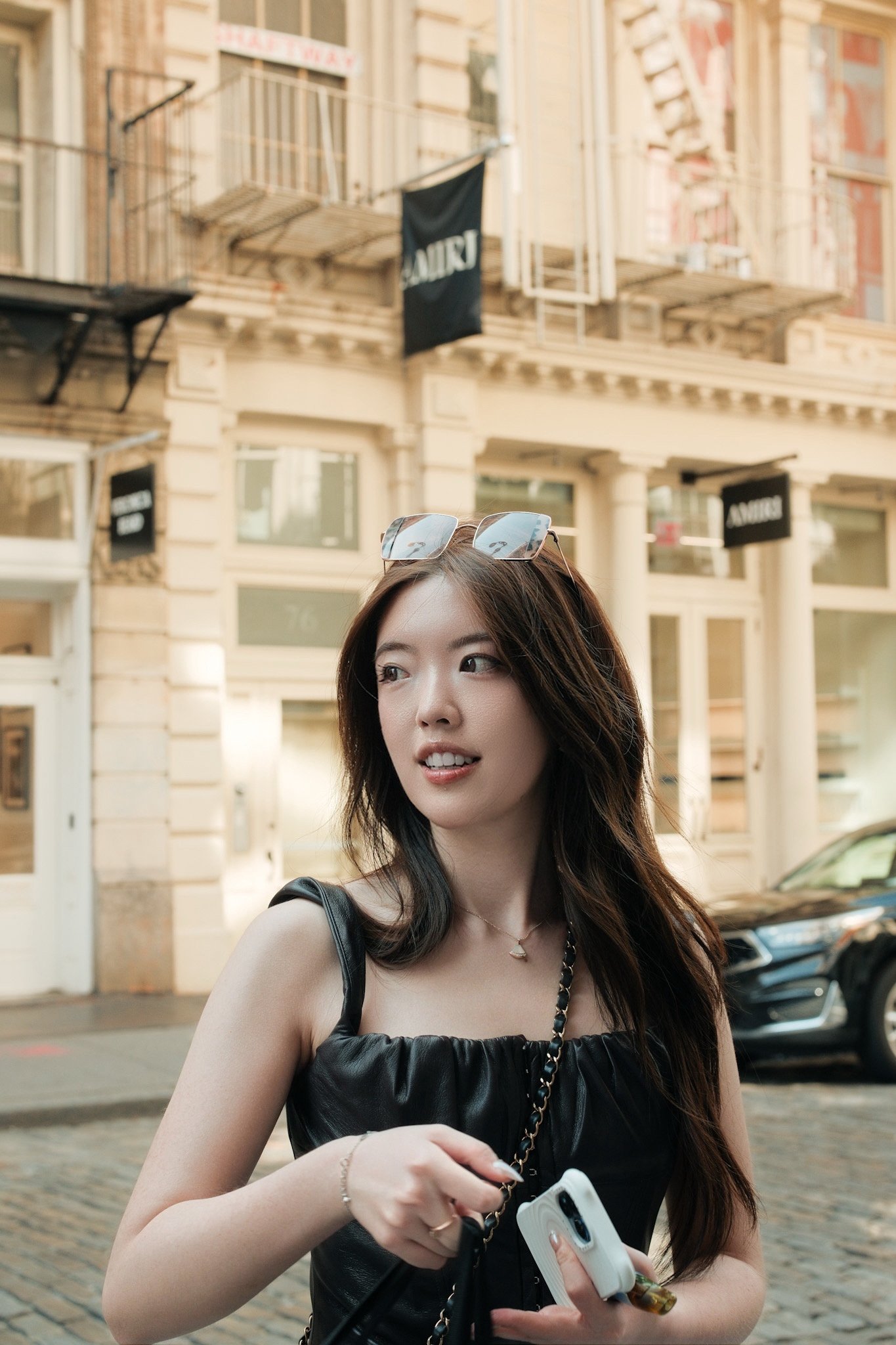 Young woman with long dark hair wearing sunglasses on her head, a black dress, and holding a white smartphone with a case and accessories, standing outdoors in front of a historic building with painted brick and black signs.