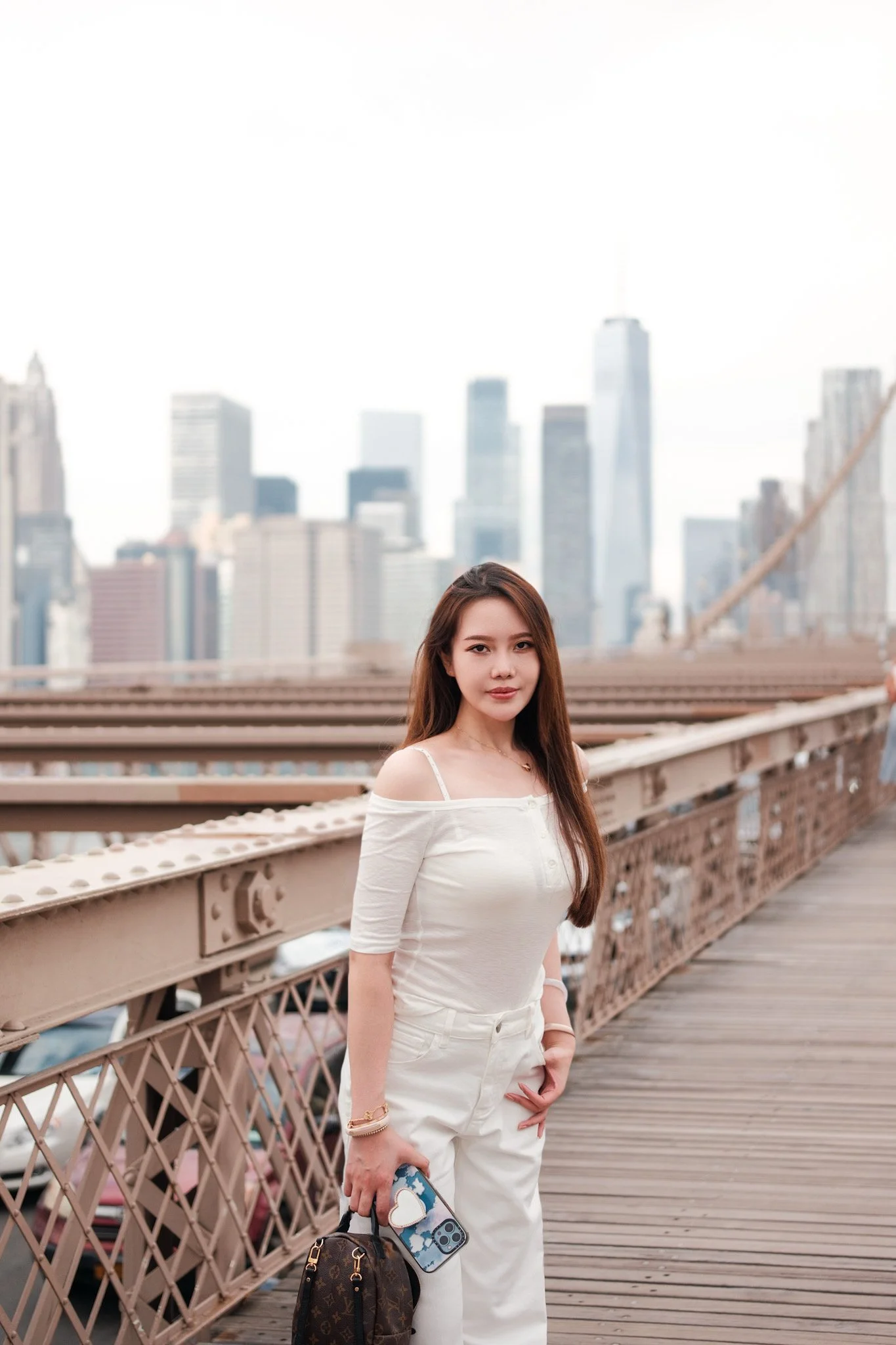 A young woman with long brown hair stands on a bridge, wearing a white off-shoulder top and white pants, holding a brown designer bag and a phone with a colorful case, with a city skyline in the background.