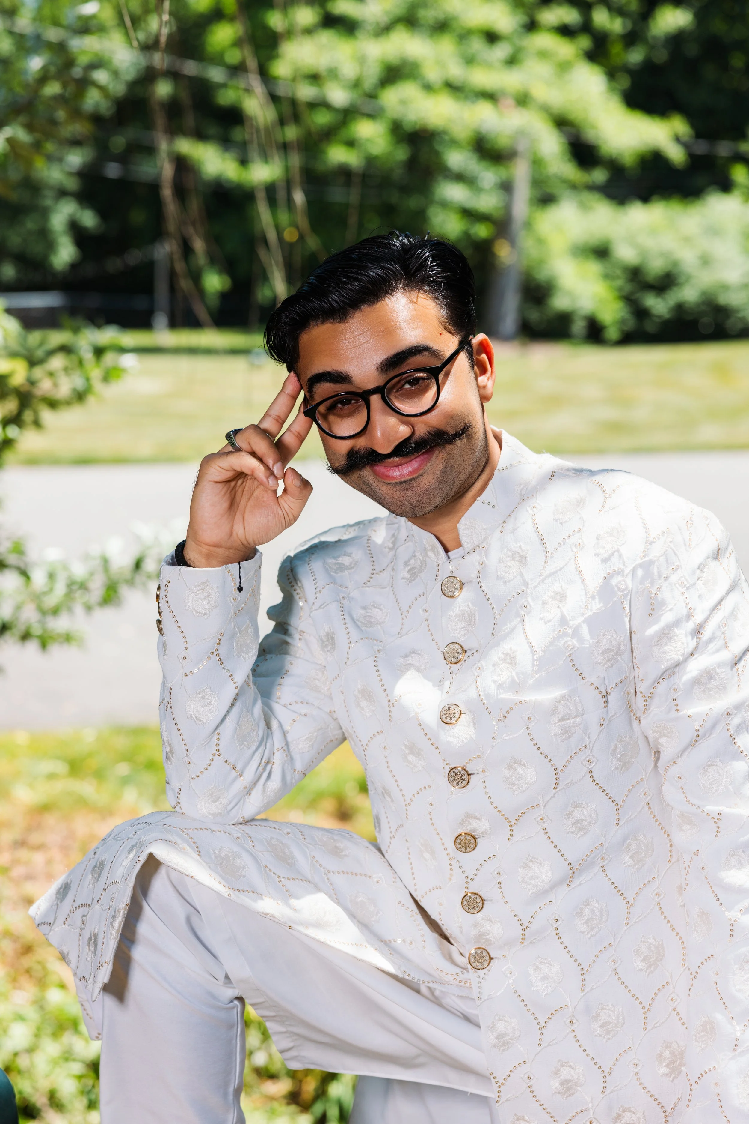 A man with black hair, glasses, and a mustache in traditional white Indian attire, sitting outdoors on a sunny day with a green, leafy background. He is smiling gently and touching his temple with his right hand.