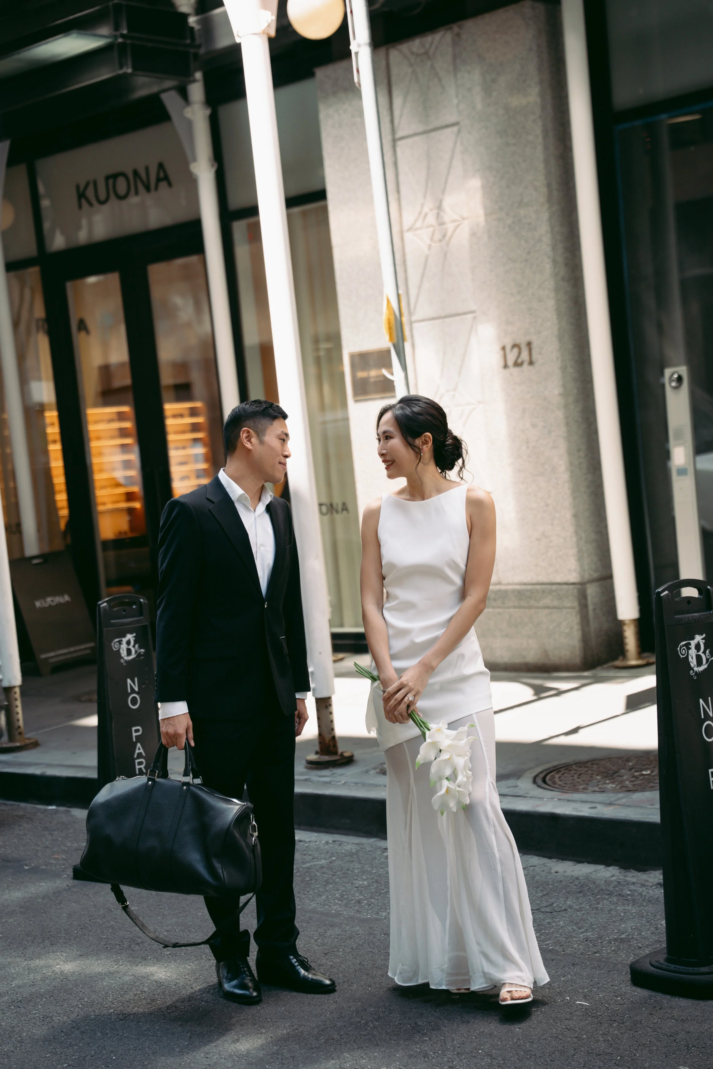 A man and woman dressed in formal attire standing and conversing on a city sidewalk in front of a building labeled 'KUONA.' The woman is holding a bouquet of white lilies. The man is carrying a black duffel bag and a black leather briefcase. They are