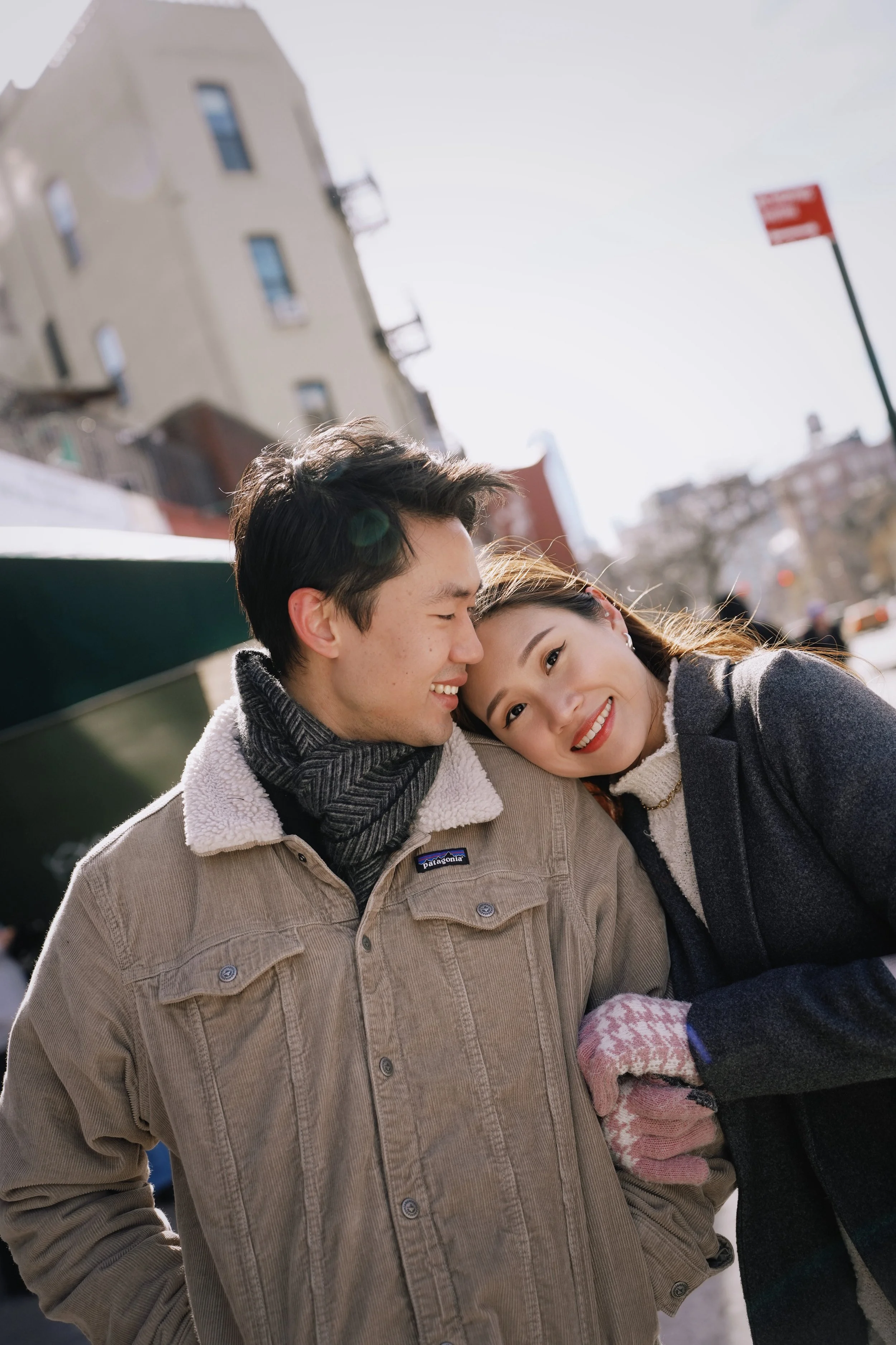 A young couple outdoors in winter, smiling with heads close together, wearing warm jackets and knit gloves, in a city setting with buildings and a street sign visible in the background.