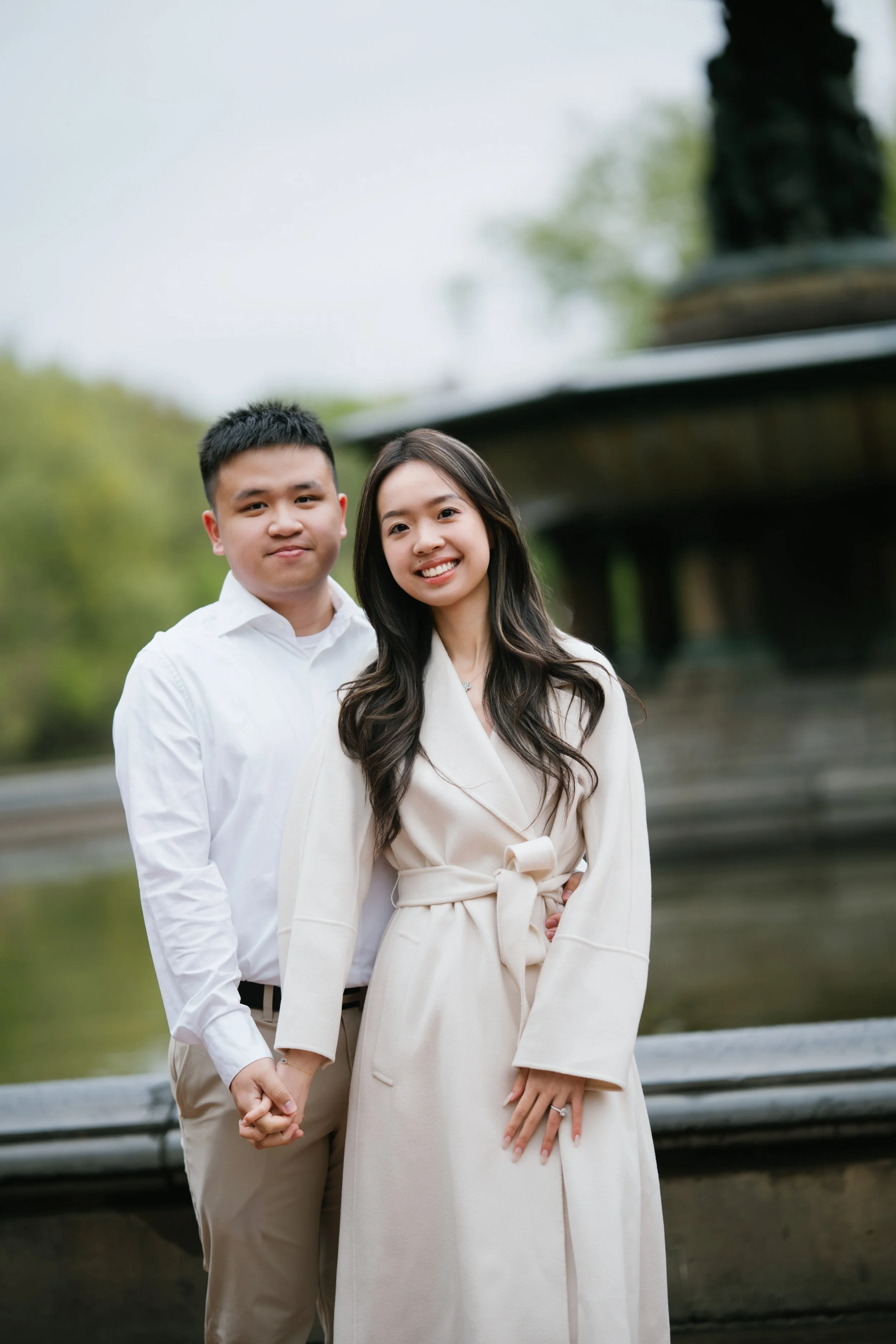 A young couple holding hands, standing outdoors near a wooden fence and a pond with trees in the background. The man has short dark hair and wears a white shirt and beige pants. The woman has long dark hair and wears a cream-colored coat with a tied 