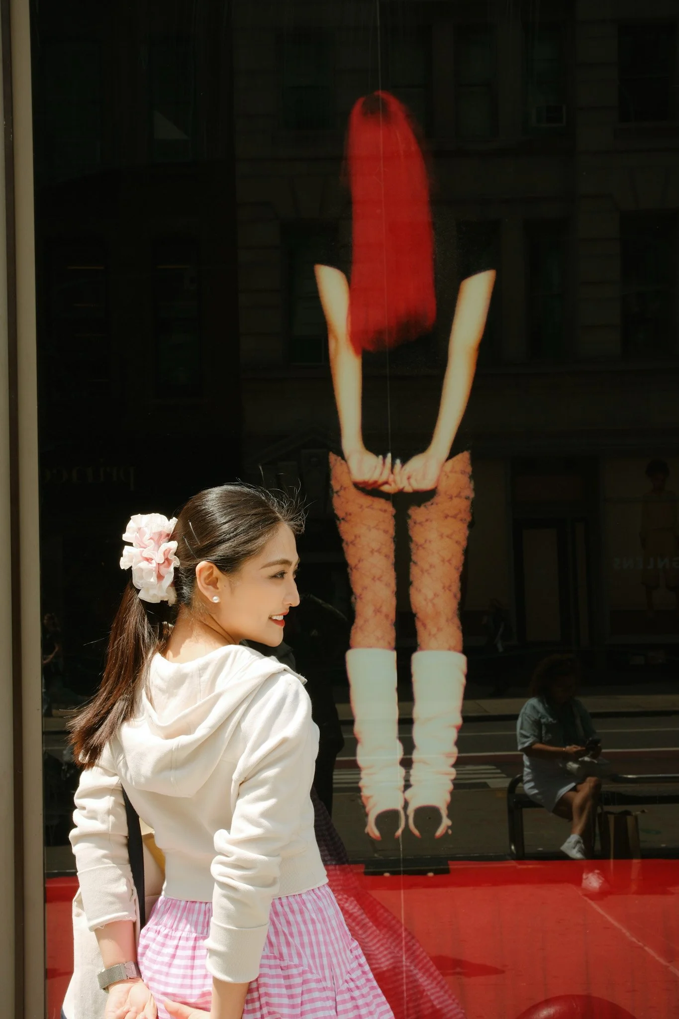 Young woman with a pink and white striped skirt, white hoodie, and a large pink scrunchie in her hair, standing outside in front of a glass window with a portrait of a woman with long red hair, black top, floral pants, and white boots reflected in it
