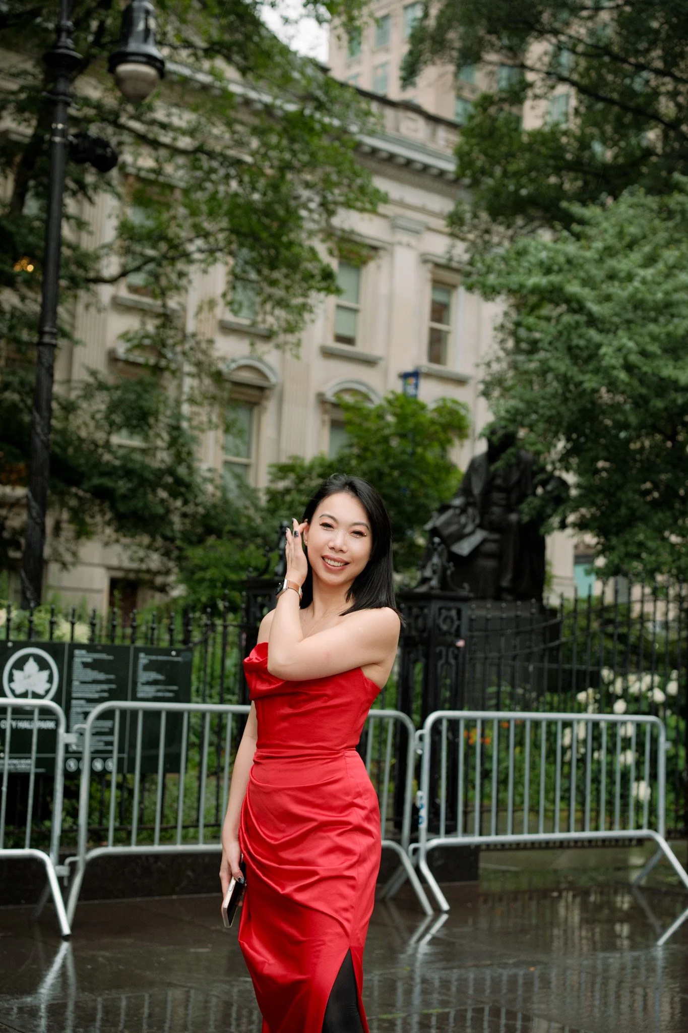 A woman in a red strapless dress smiling and touching her hair while standing outdoors near metal barriers in a park with trees and historic buildings in the background.