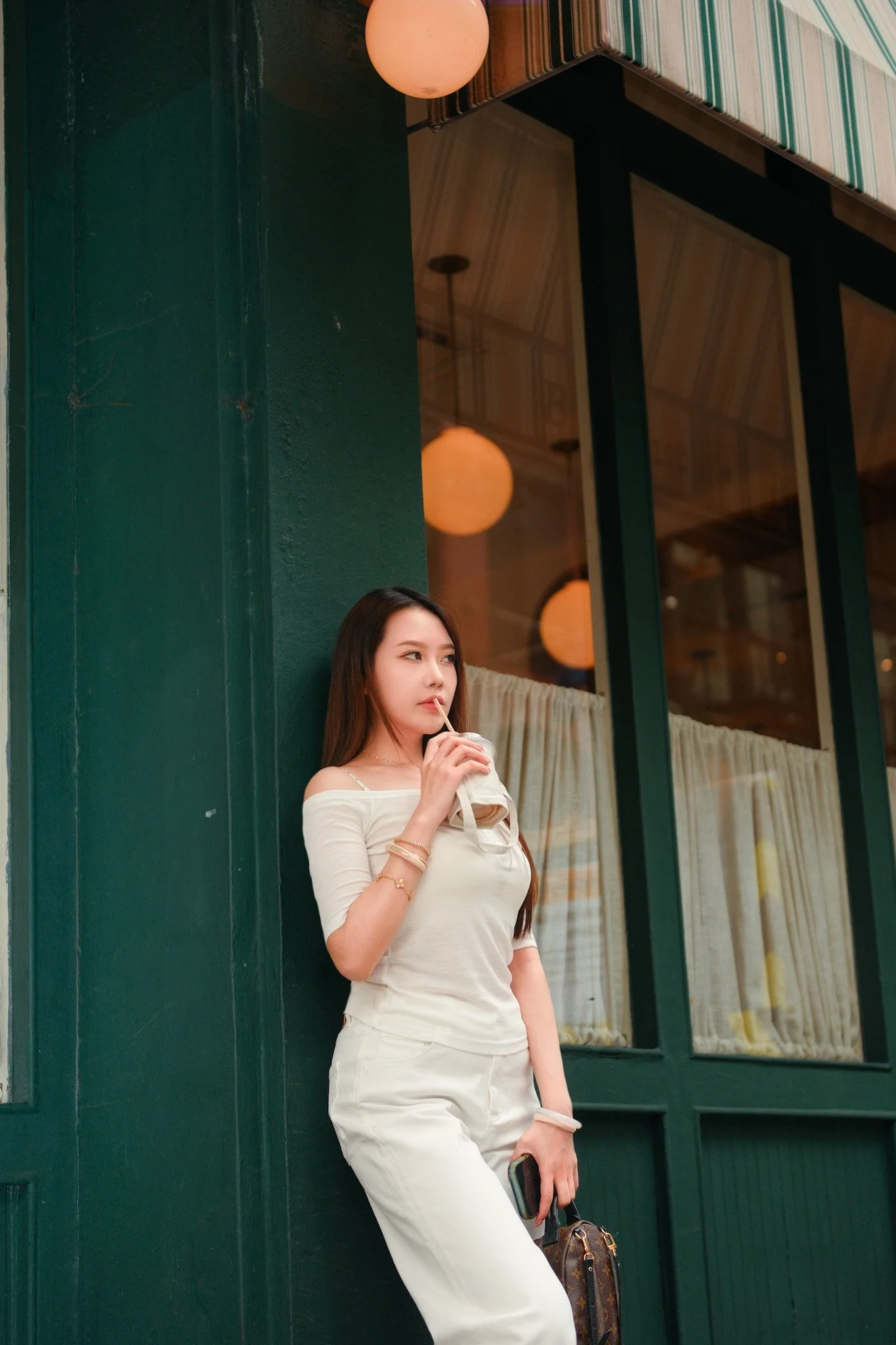 A young woman with long dark hair wearing a white off-shoulder top and white pants, standing against a dark green wall outside of a building with large glass windows and a striped awning, holding a beverage with a straw in her right hand and a handba