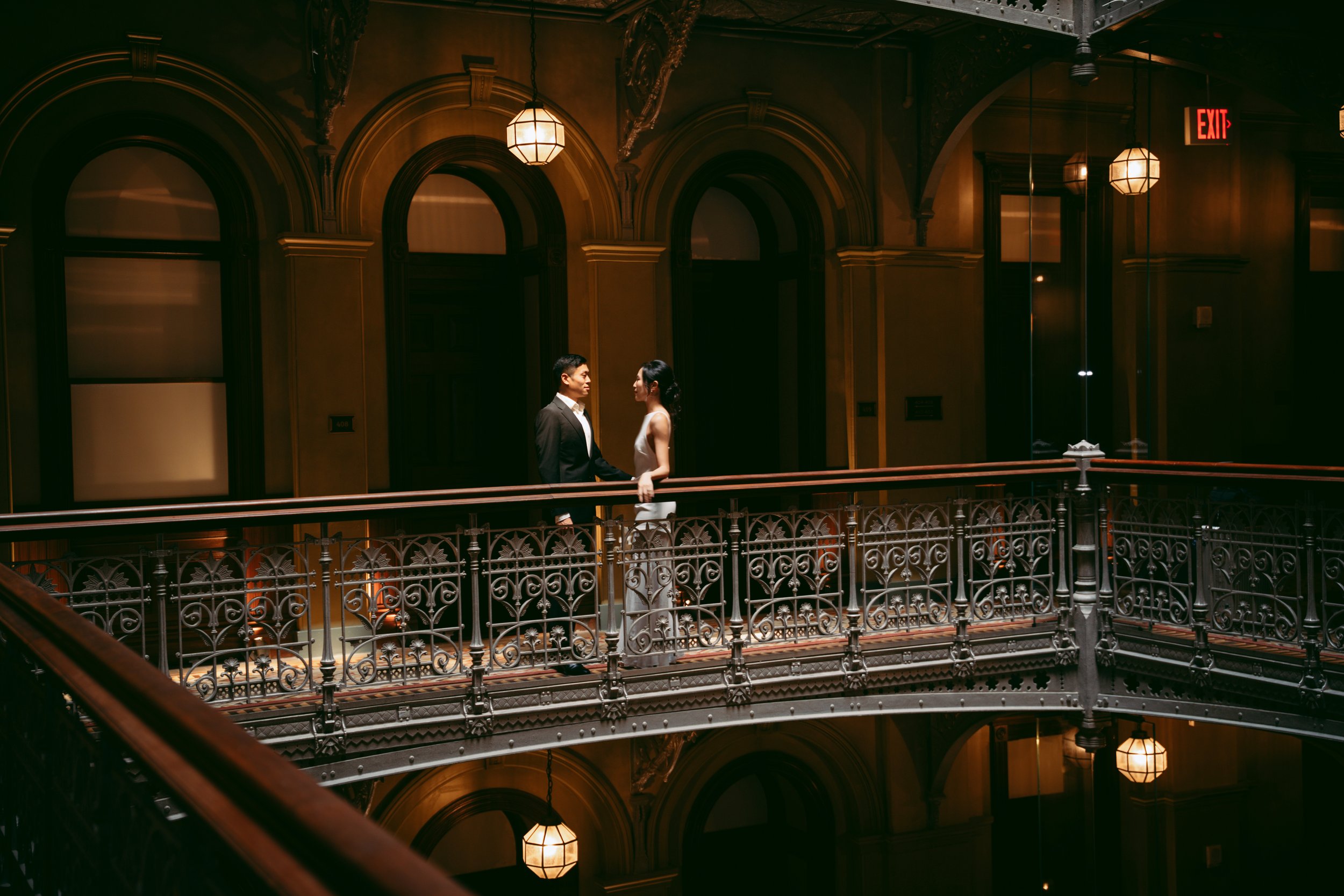 A man and woman standing close on a balcony inside a dimly lit, ornate building with arched windows and decorative iron railing, holding hands and looking at each other.