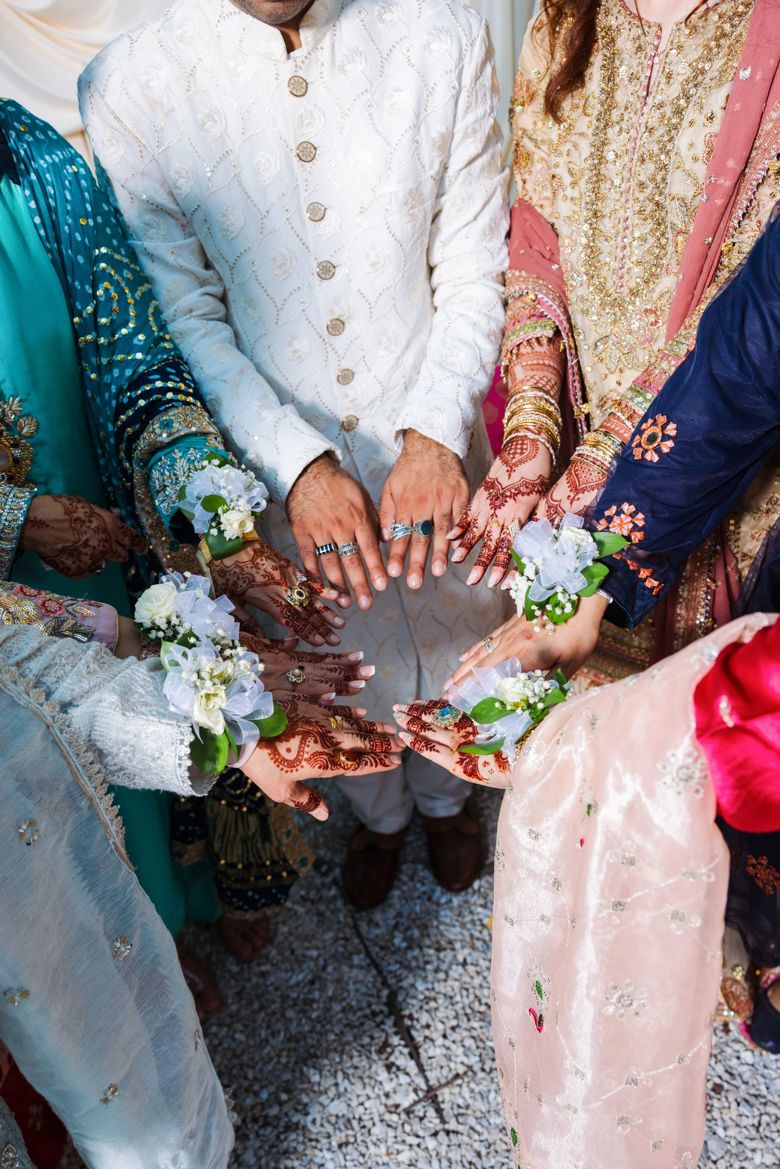 Group of people wearing traditional, colorful Indian wedding attire, showing their hands with rings and henna designs.
