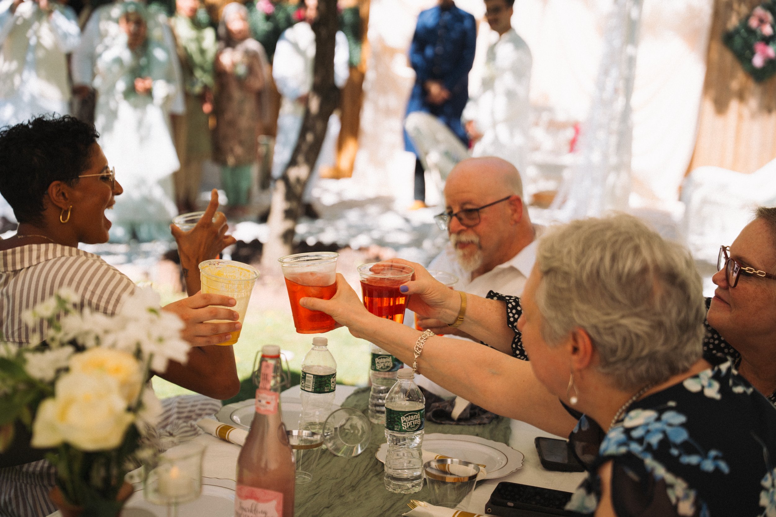 People at a celebration raising drinks in a toast, seated around a table outdoors with floral decorations and bottles of drinks.