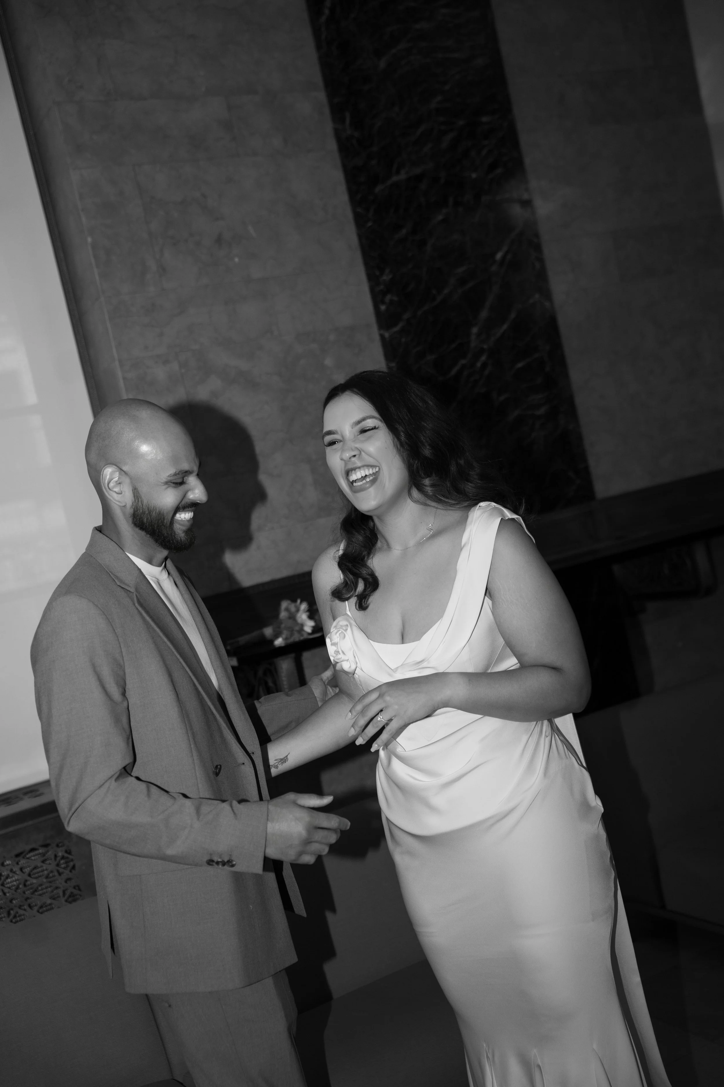 A man and woman are dancing and smiling at each other in an indoor setting, both dressed in formal attire.