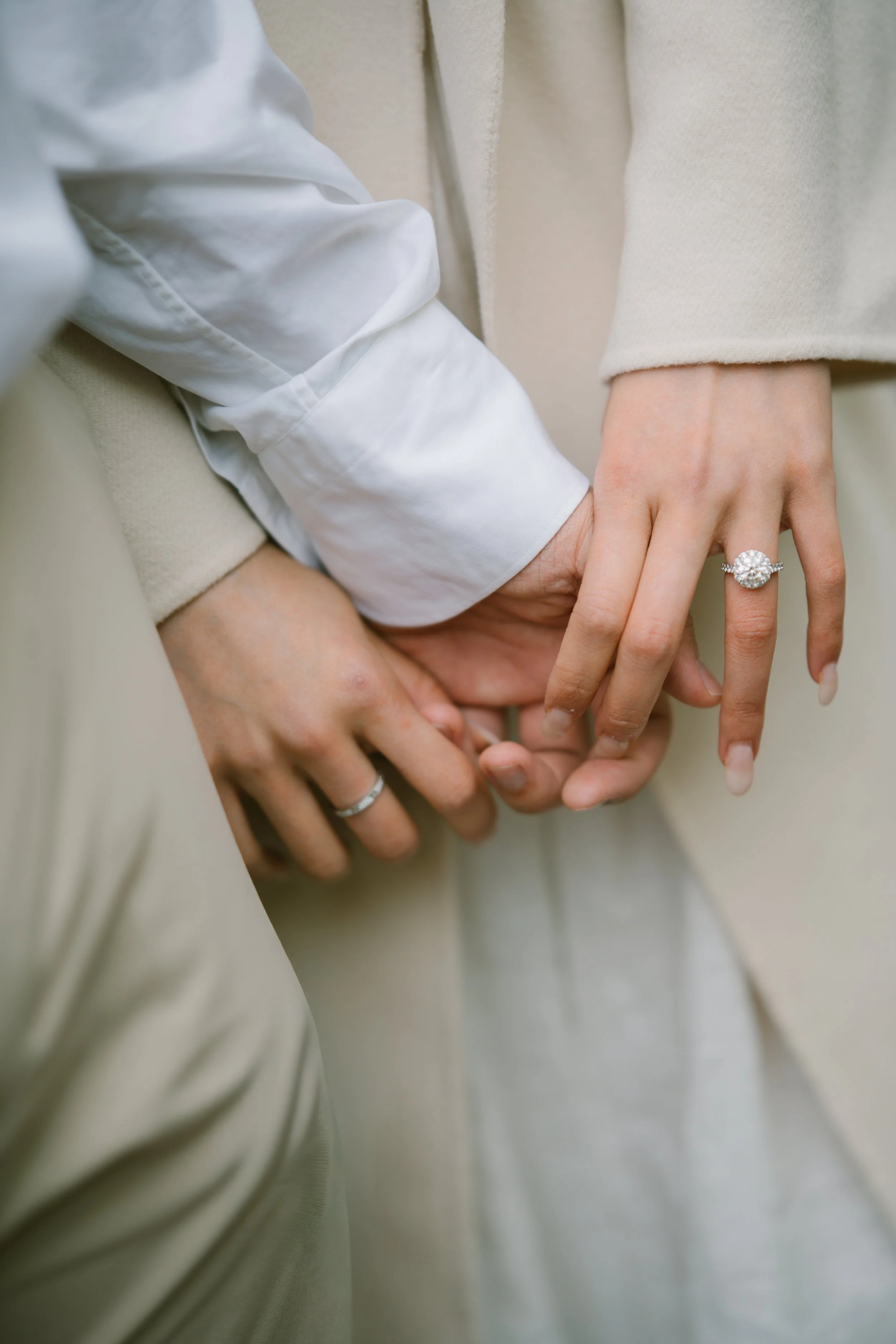 Close-up of two hands holding each other, one with a diamond engagement ring, dressed in neutral-colored clothing.