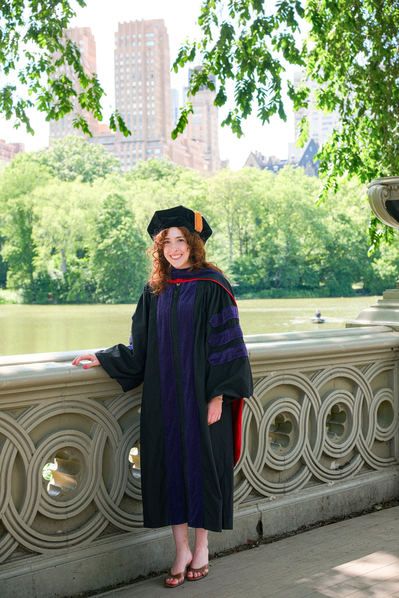 A woman in a graduation gown and cap smiling outdoors on a bridge with green trees and tall buildings in the background.