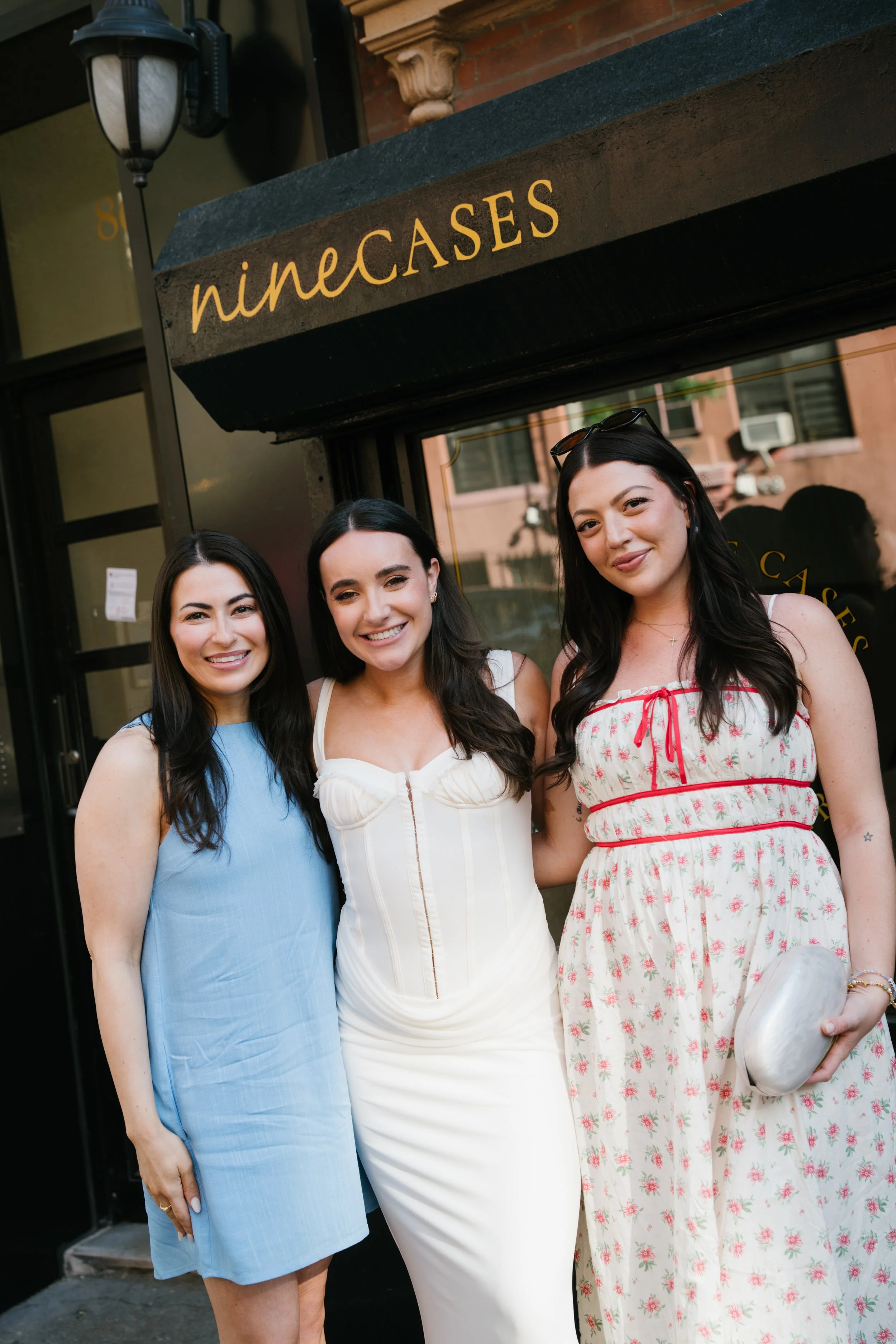 Three women standing together outside a building with a sign that reads 'ninecases' above their heads.