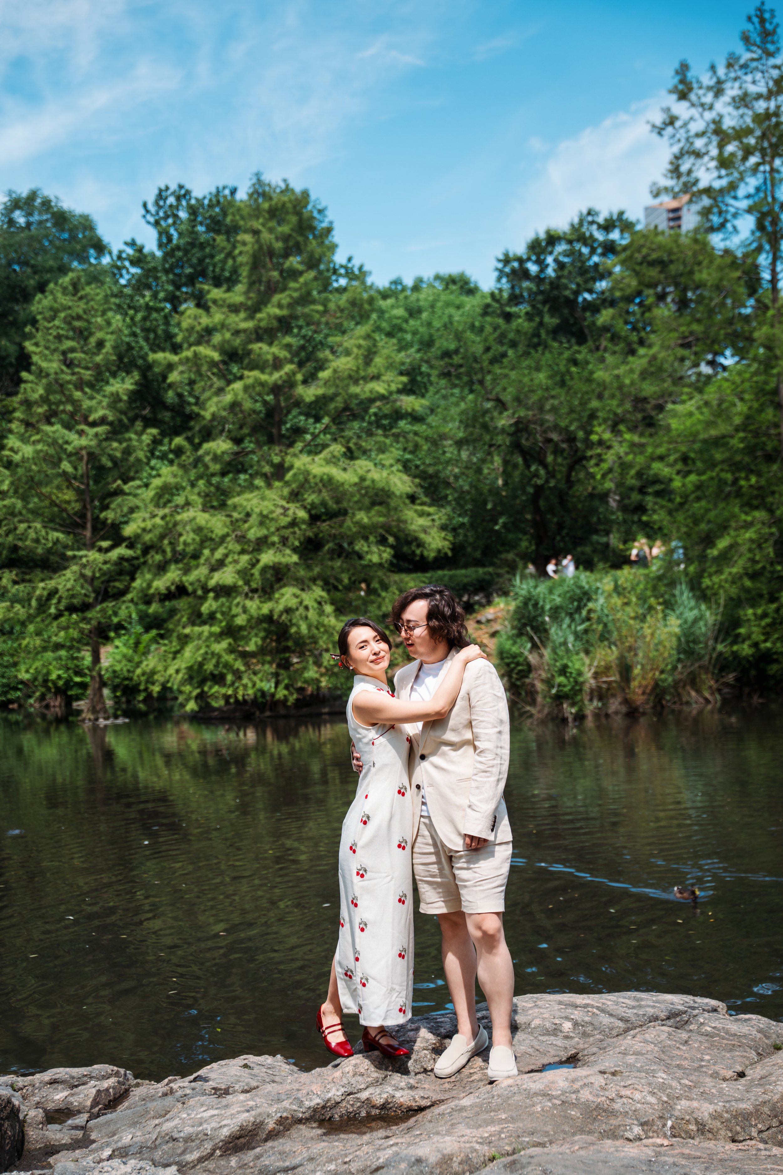 A couple standing on rocks by a lake, surrounded by green trees, with a clear blue sky overhead.
