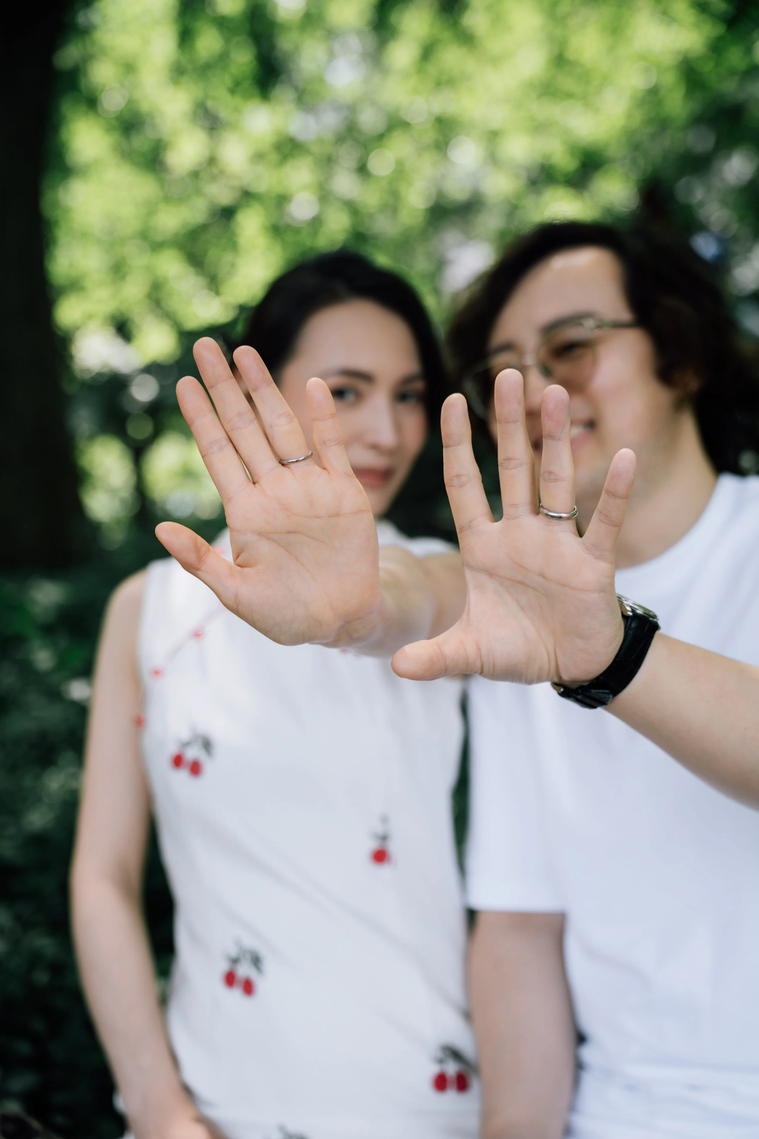 Two women wearing white dresses with red and green cherry prints, standing outdoors with a blurred background of green trees, showing their hands with silver rings raised towards the camera.