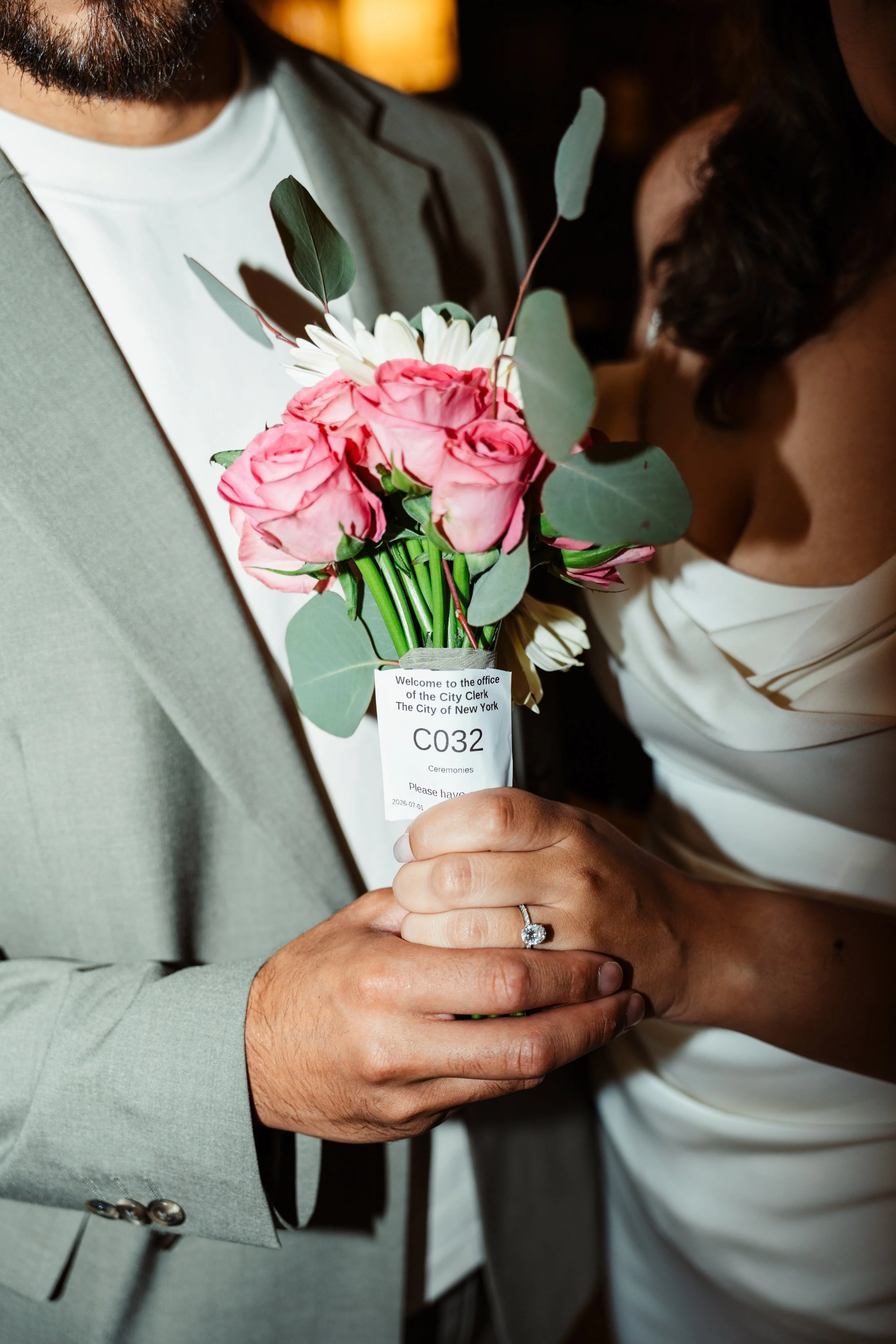 Close-up of a couple holding hands, with the woman's hand displaying a diamond ring, and a bouquet of pink roses and white flowers resting on their hands. The couple is dressed formally, and the bouquet has a tag from the City of New York.
