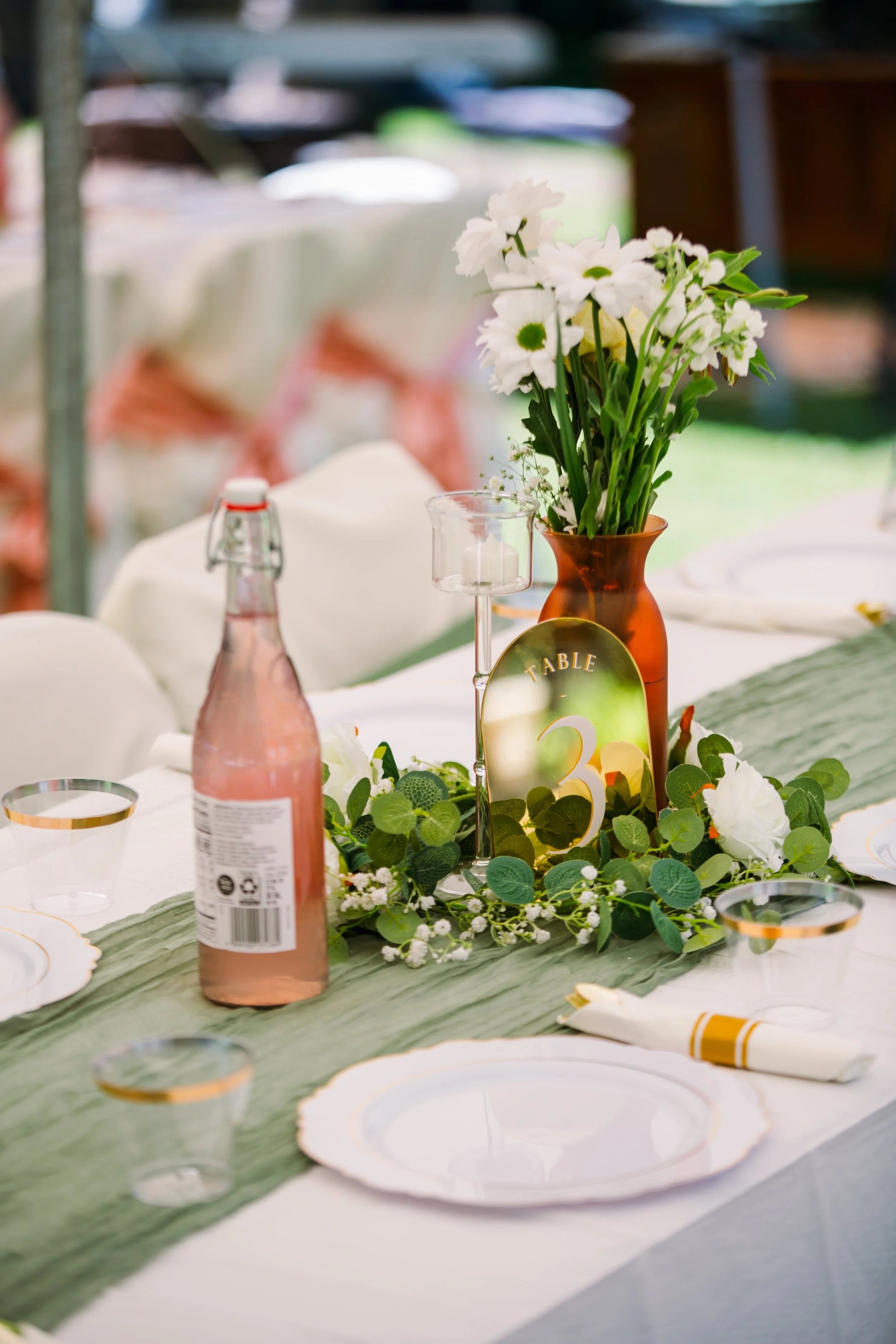 Table decorated with a green table runner, white plates with gold rims, floral centerpiece of white flowers in a brown vase, greenery, and a table number sign.