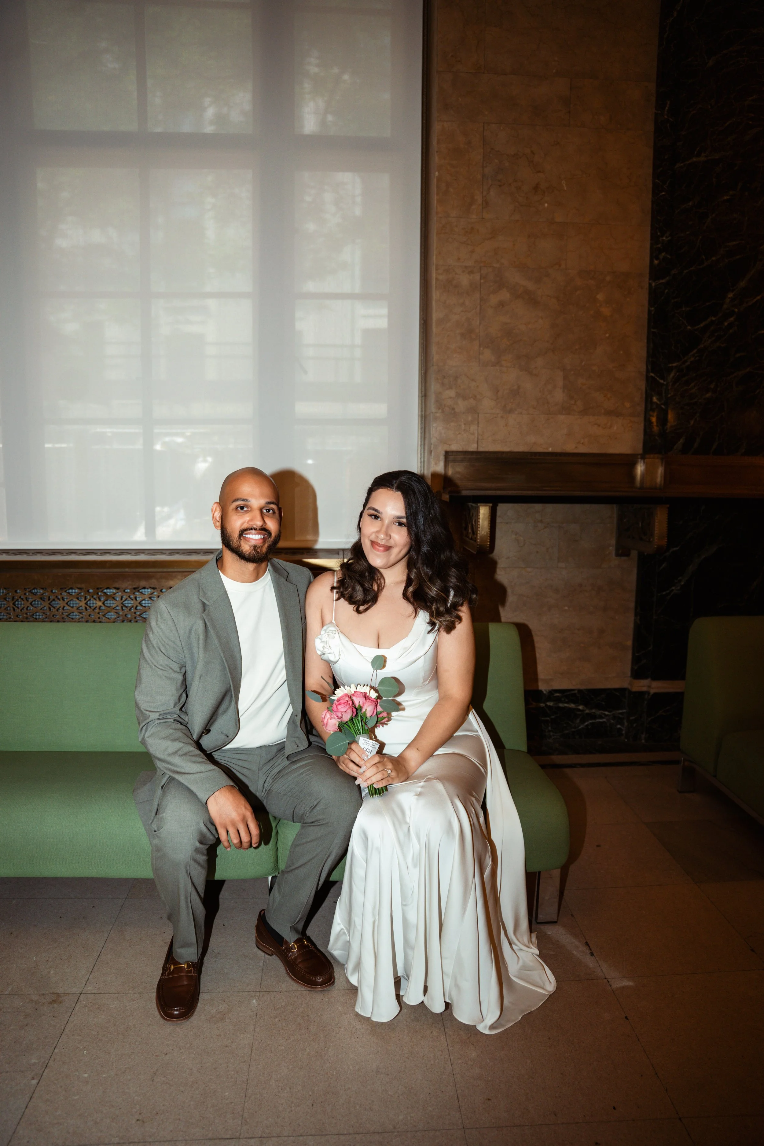 A smiling man in a gray suit and a woman in a white dress sitting on a green bench in an elegant room. The woman is holding a small bouquet of pink and white flowers.