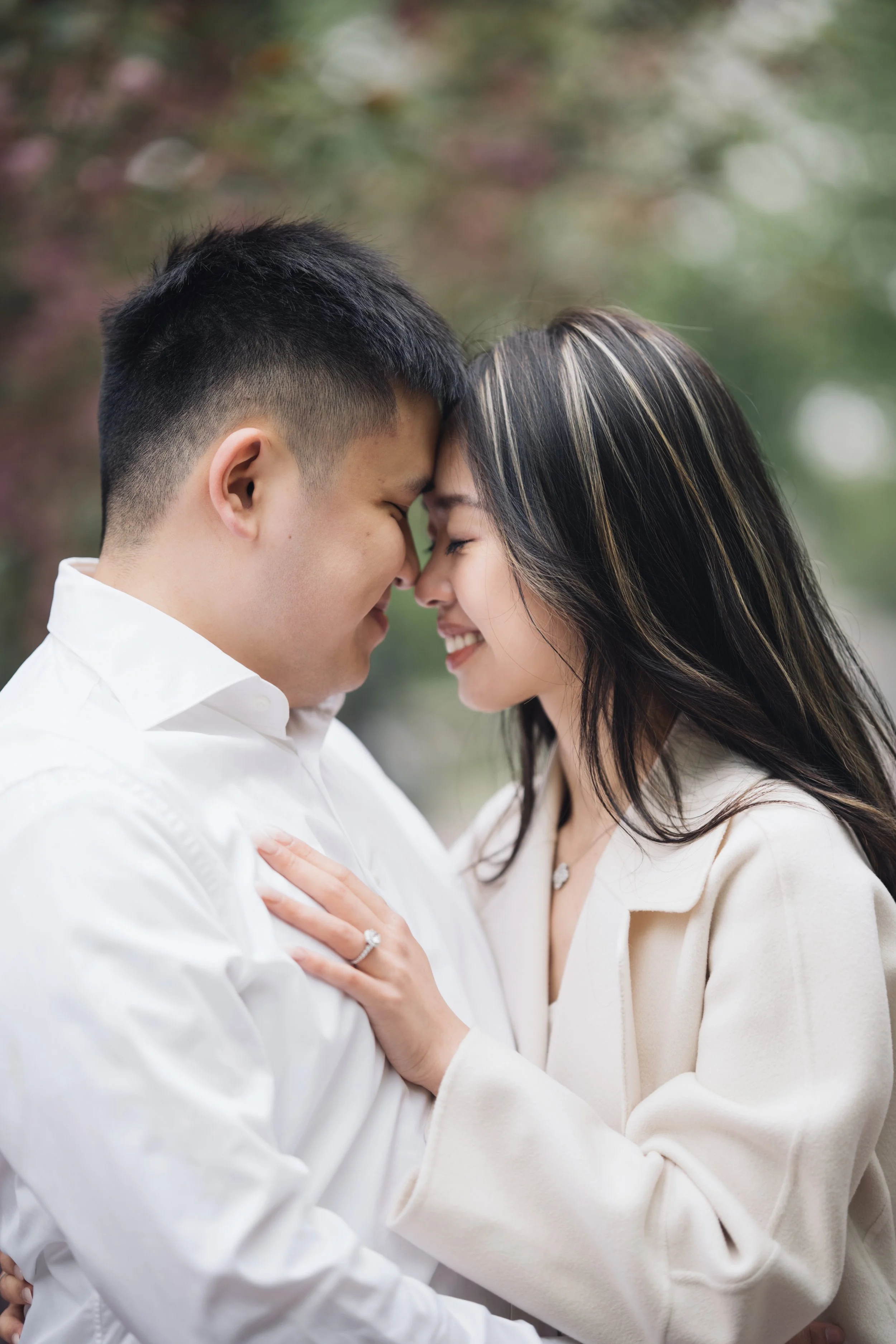 A couple sharing a close, tender moment with their foreheads pressed together, eyes closed, and gentle smiles, outdoors with blurred greenery in the background.