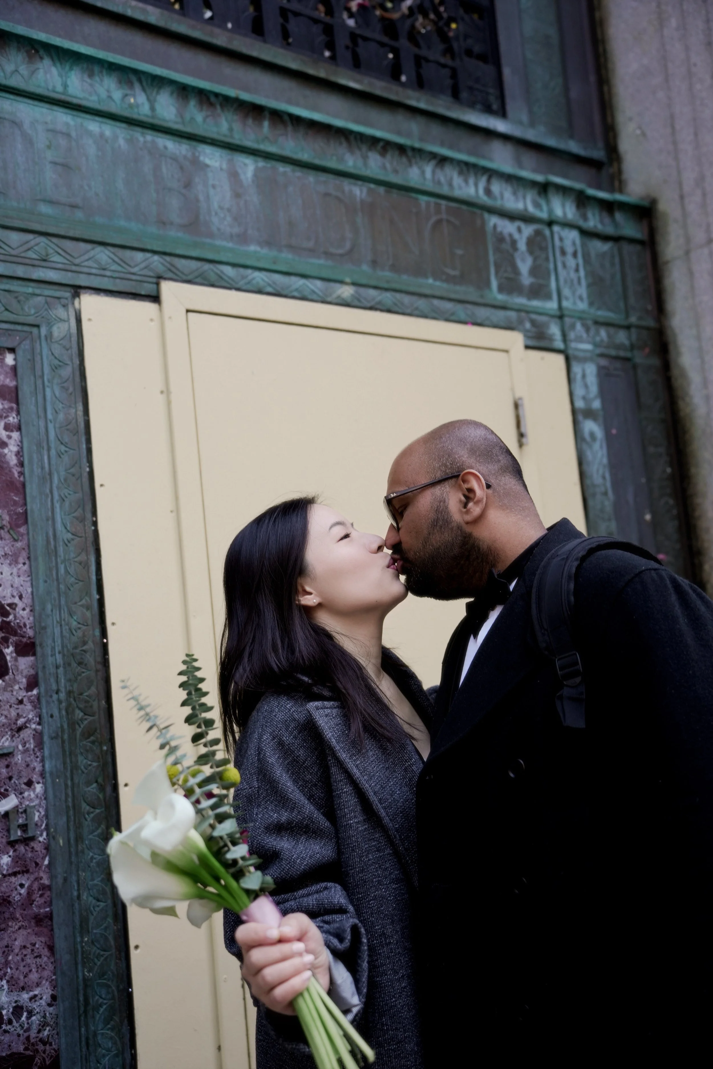 A couple is standing close together, about to kiss, in front of a decorative wall with a yellow door. The woman is holding a bouquet of white flowers in her left hand. The woman has long dark hair and is wearing a gray blazer, and the man is bald, we