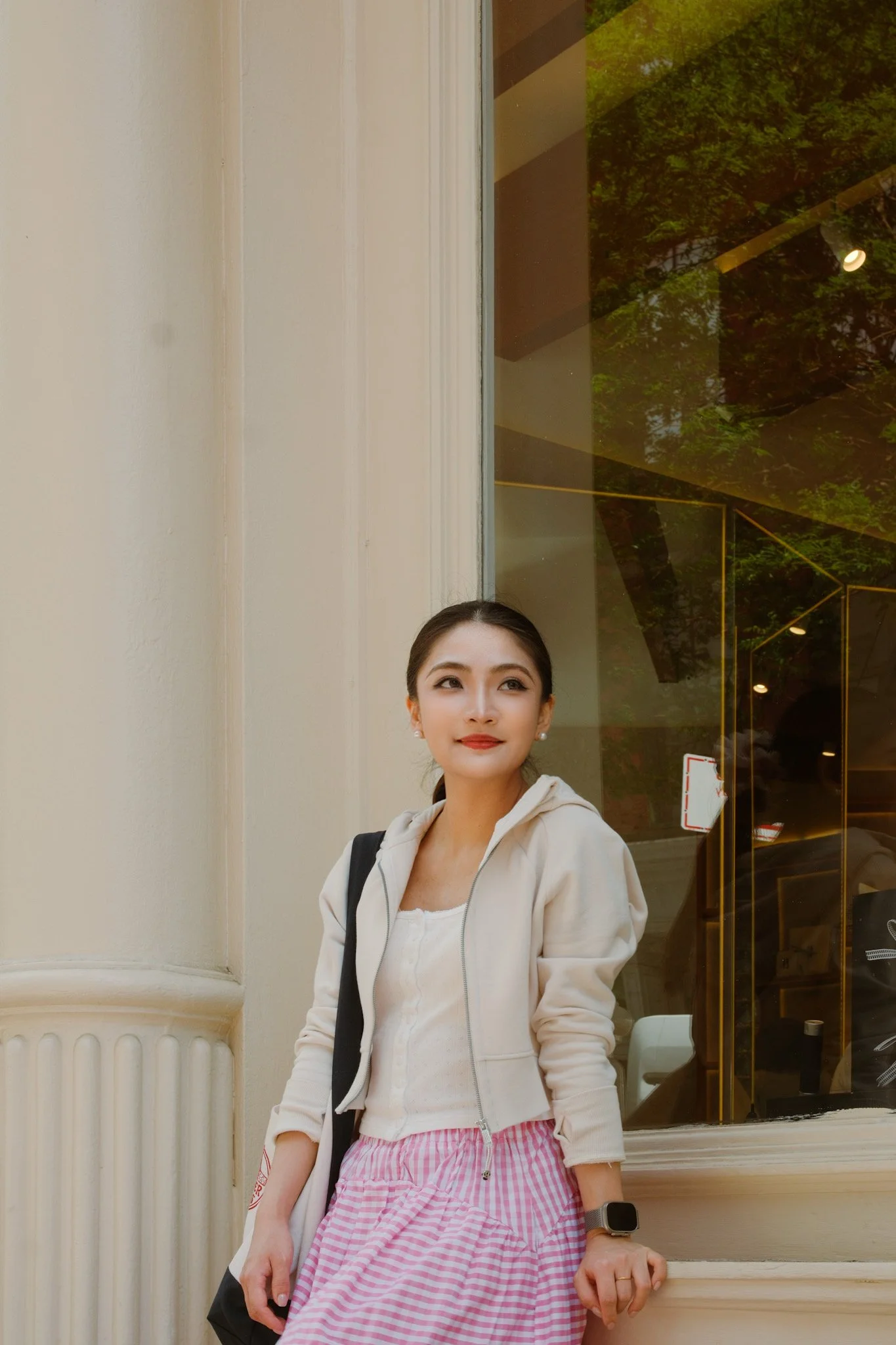 A woman with dark hair tied back, wearing a light-colored hoodie, pink and white striped skirt, and a smartwatch, standing outside a building with a glass window and reflection of green trees, looking slightly to the side with a slight smile.