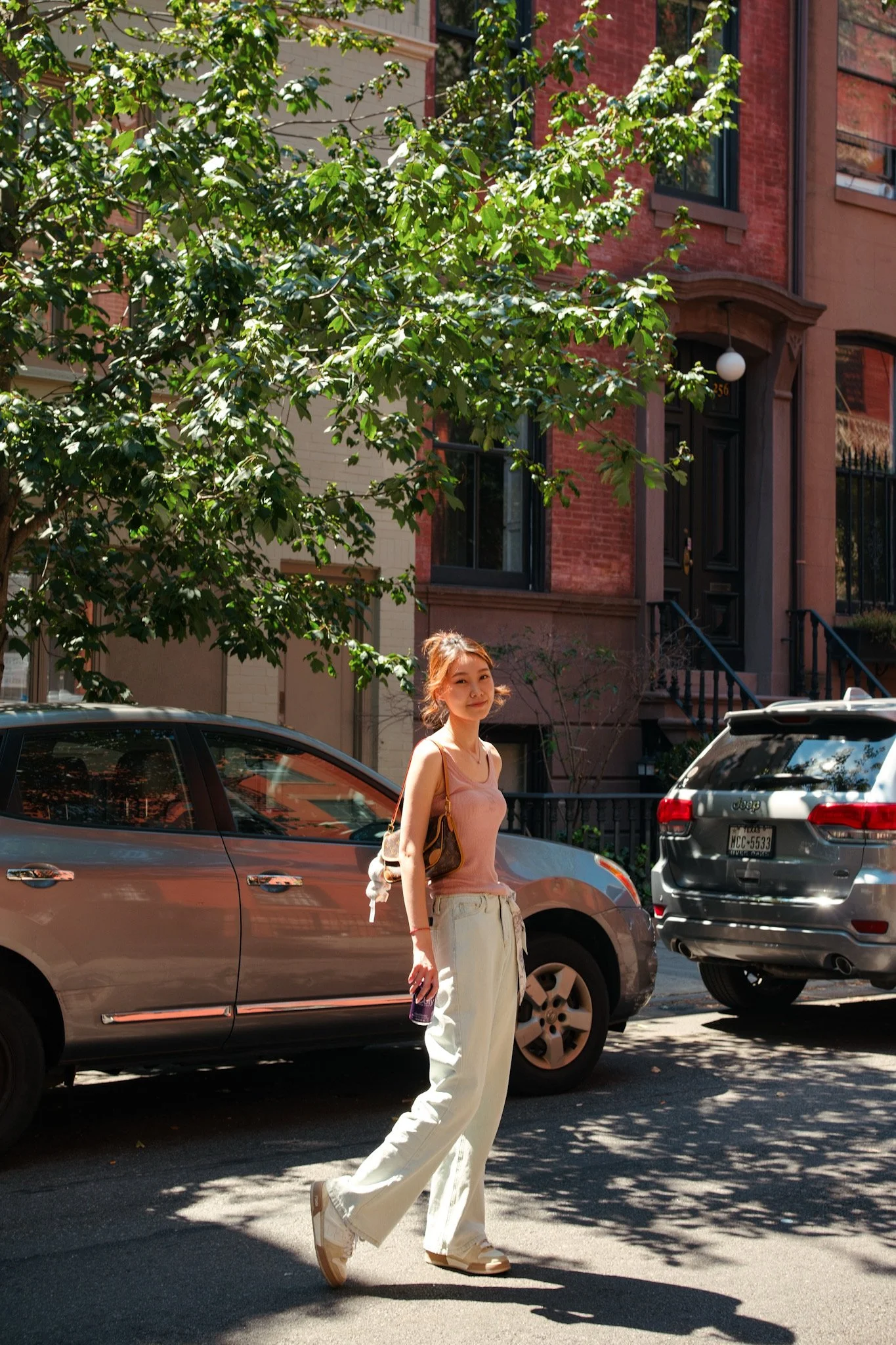 A young woman with auburn hair, wearing a pink sleeveless top and wide-leg white pants, is walking across a city street in front of parked cars and brick row houses, carrying a tote bag and holding a purple object in her right hand.