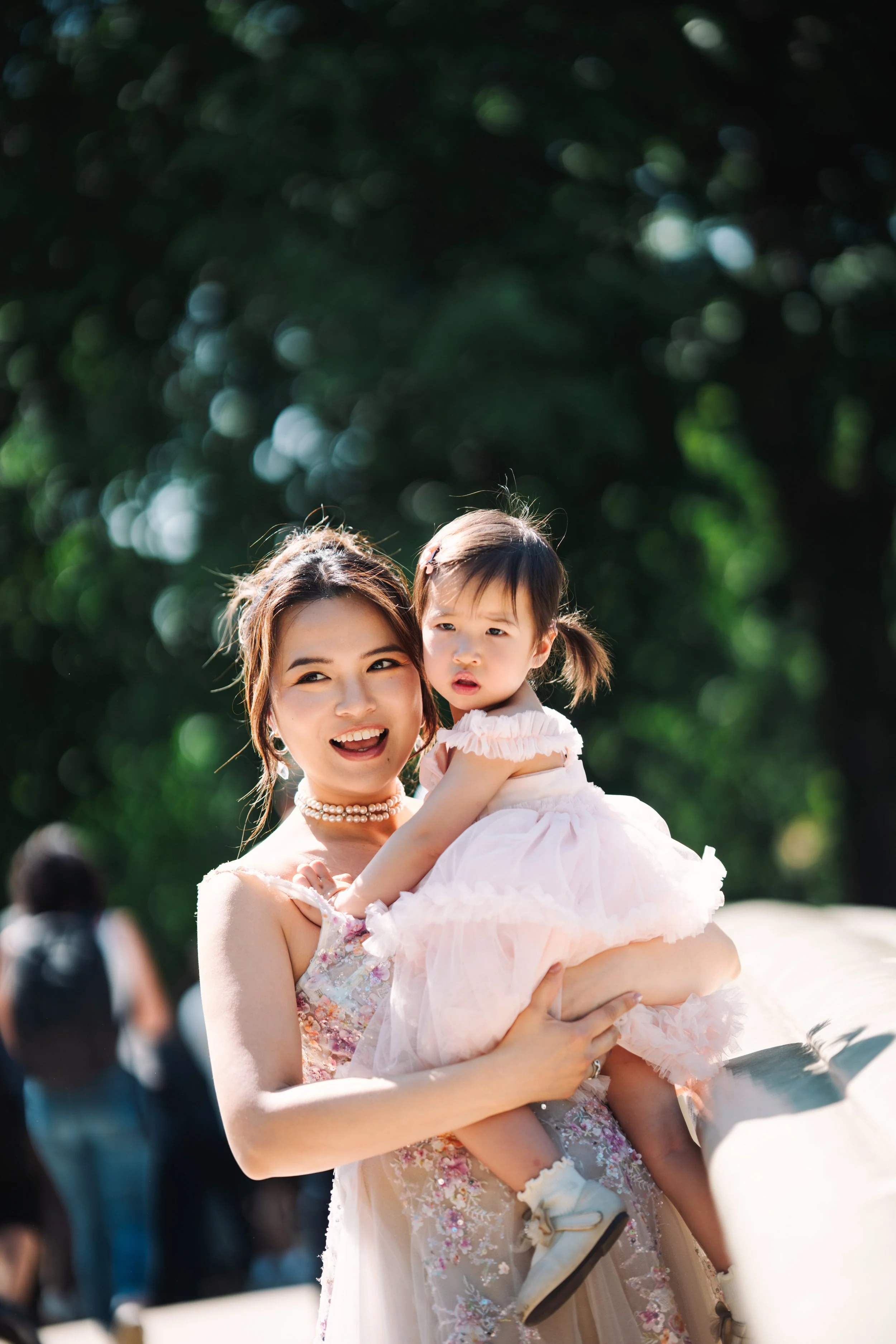 A woman holding a young girl outdoors, both smiling, with a blurred background of green trees and other people.
