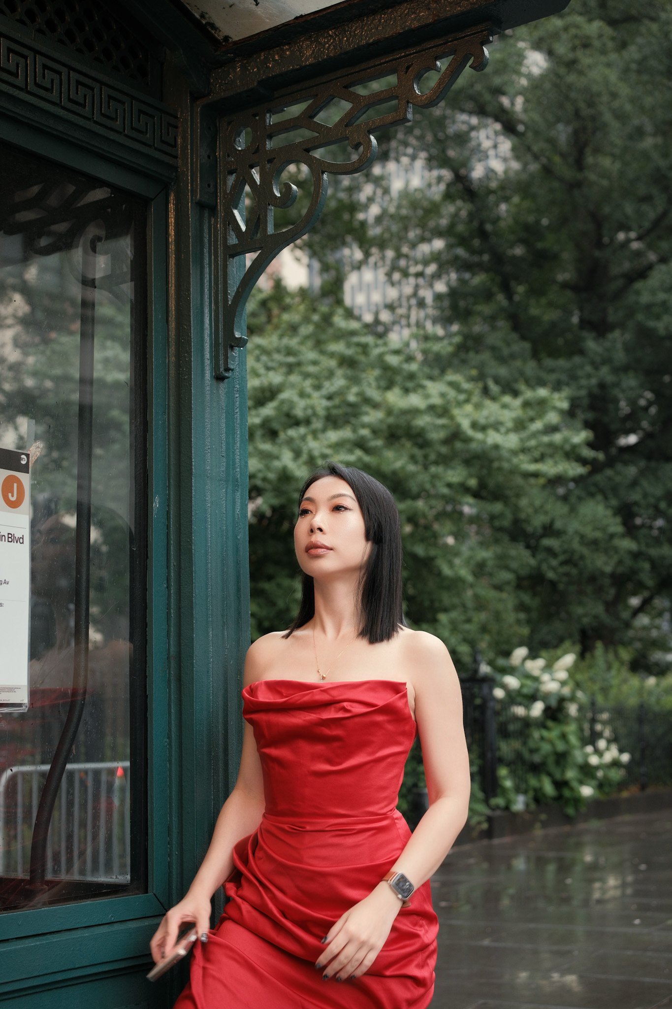 A woman wearing a red strapless dress standing outdoors near a green telephone booth with trees in the background.