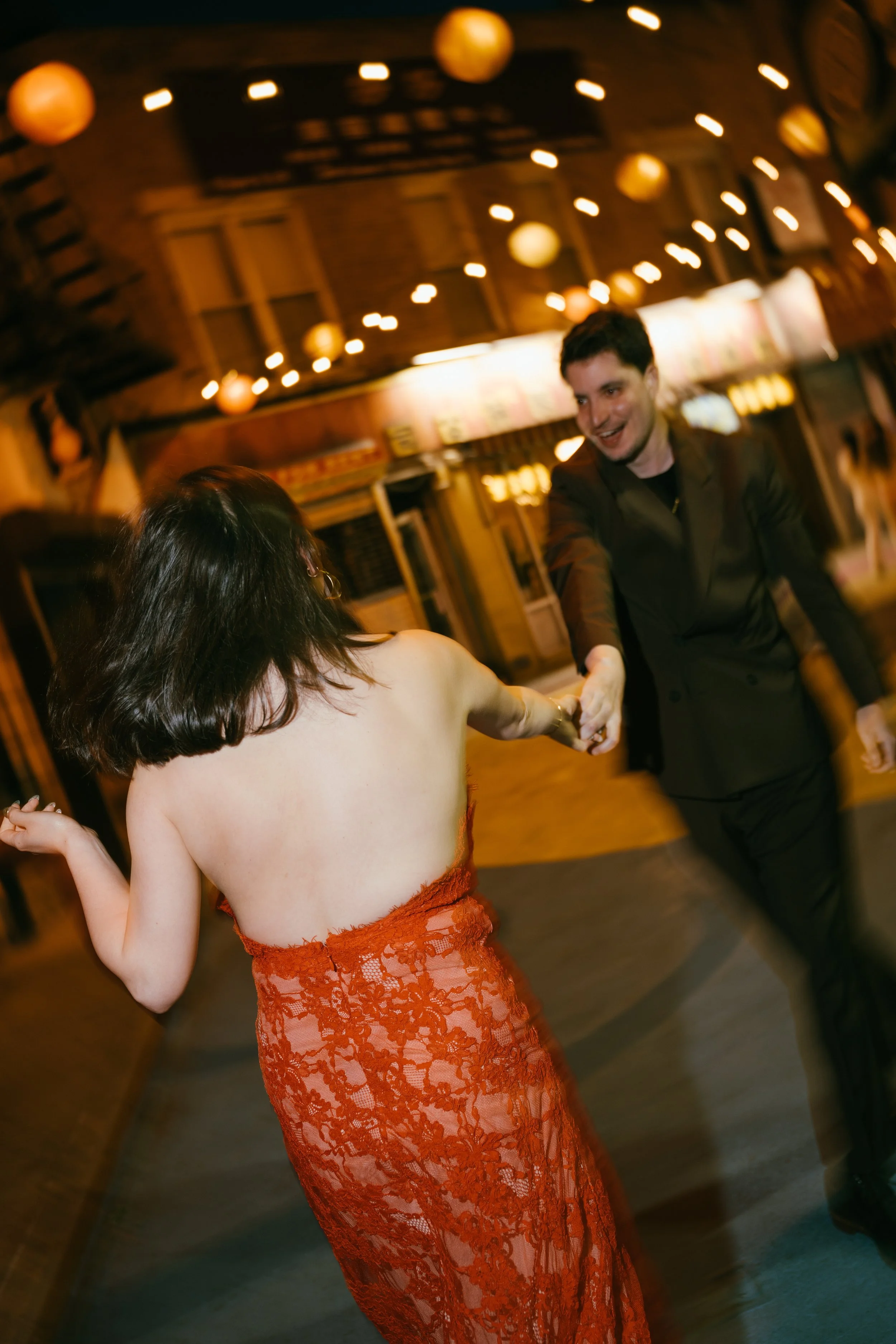 A man in a dark suit holding hands and dancing with a woman in a red lace dress at a party or celebration with string lights hanging overhead.