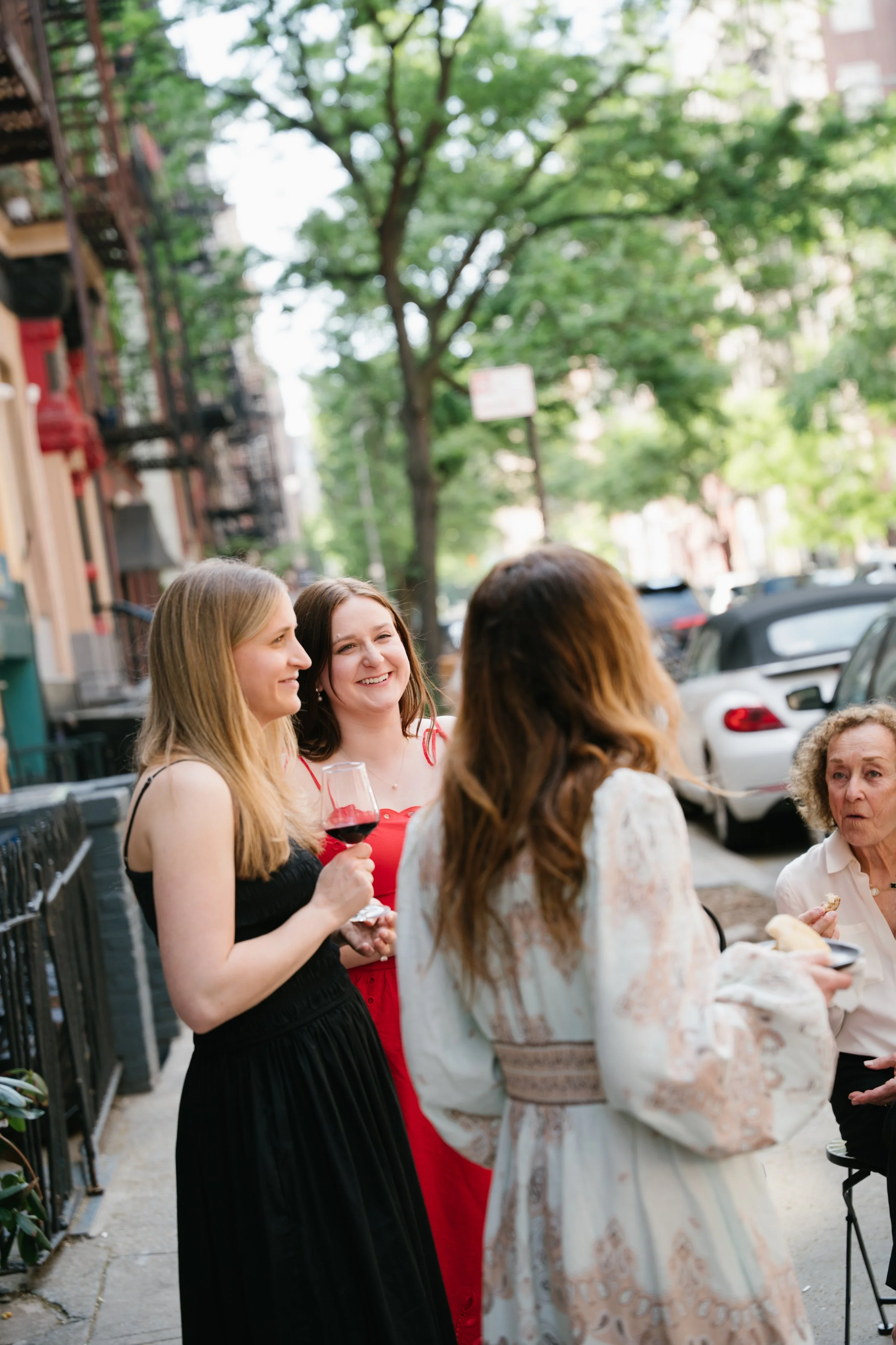A group of women standing and talking on a city sidewalk, with buildings, parked cars, trees, and street signs in the background.