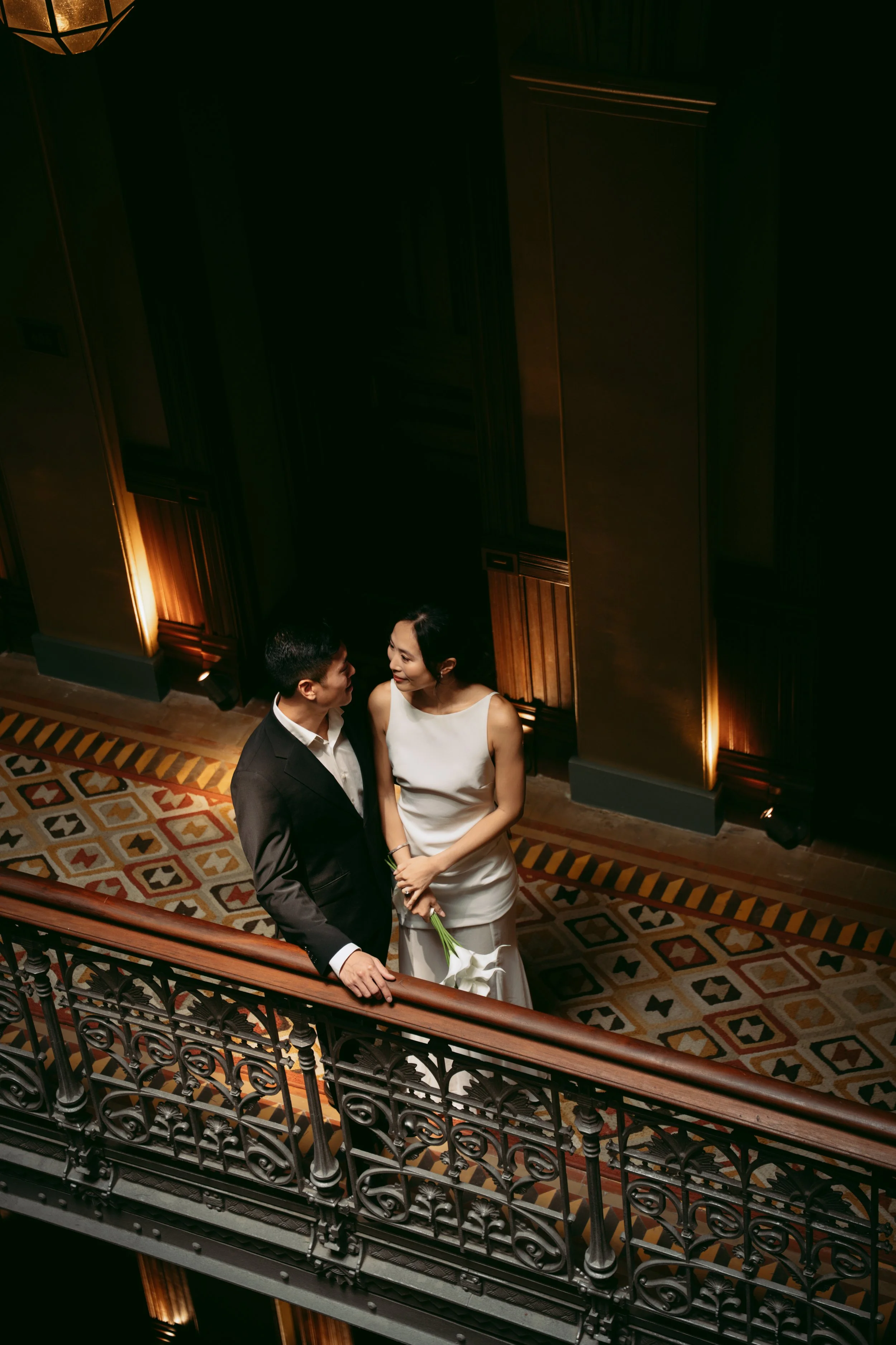 A man and woman stand on a staircase in a dimly lit, elegant setting. The man is dressed in a black suit and white shirt, the woman in a white dress holding white flowers. They are gazing at each other.
