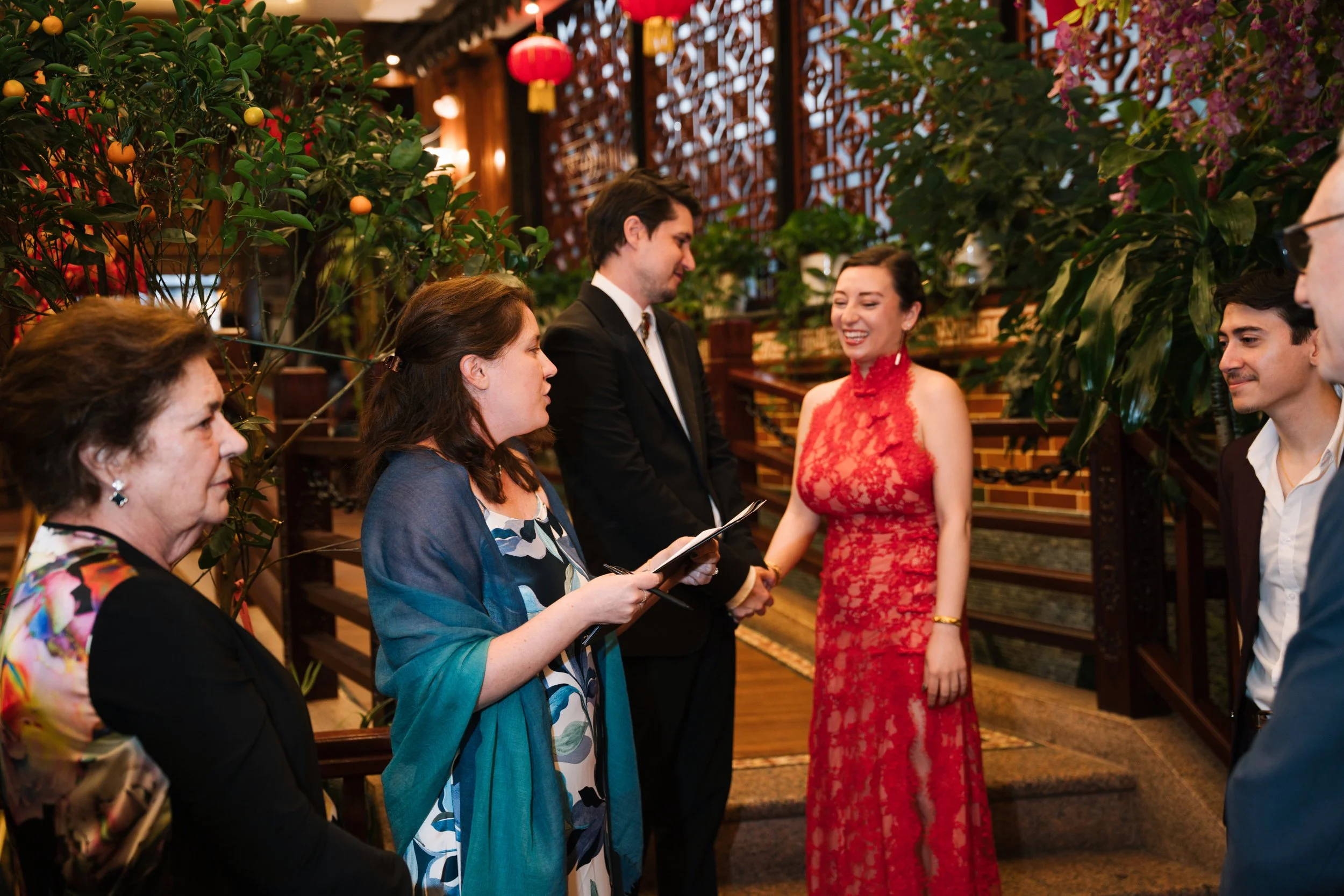 A group of people at a social gathering, with some people talking and smiling. The setting appears to be a decorated indoor space with plants, lanterns, and ornate wooden panels.
