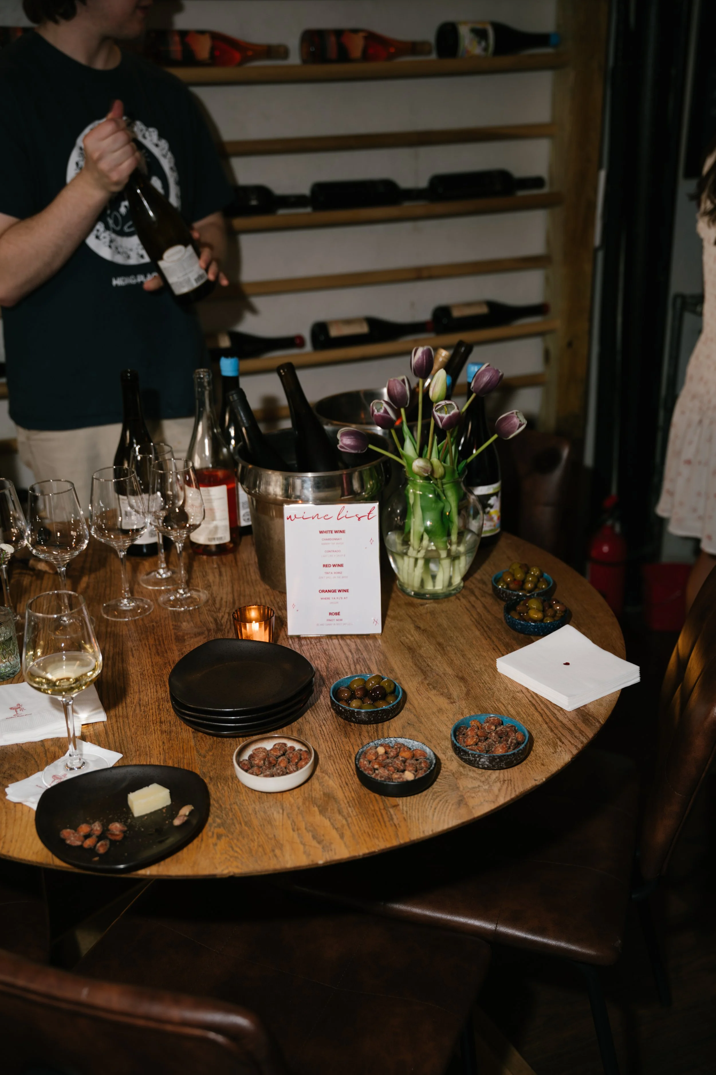 A dining table with wine bottles, glasses, bowls of nuts and snacks, a vase with tulips, and a wine list menu inside a cozy room