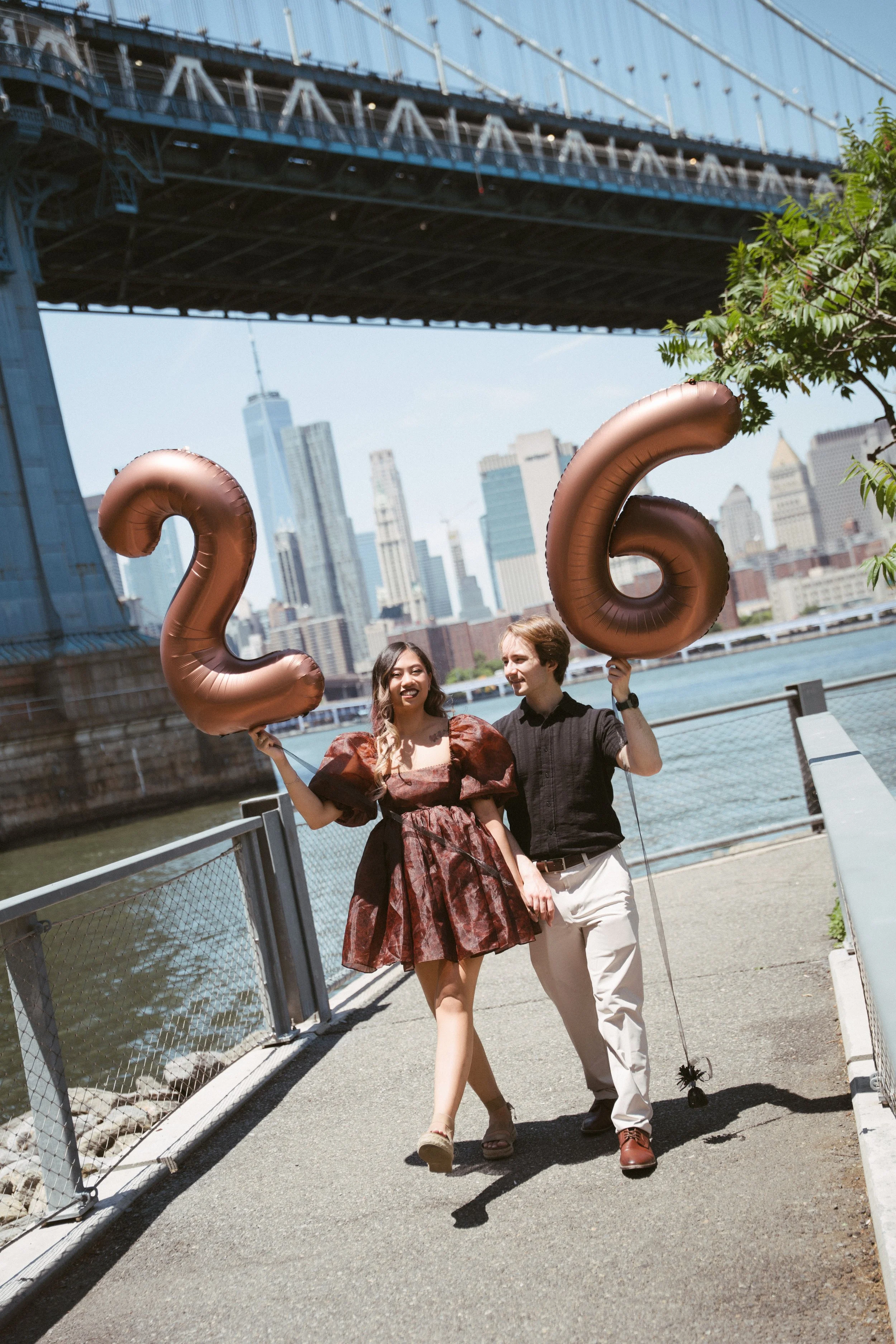 A couple walking along a waterfront pathway under a bridge in New York City, holding large bronze-colored balloons shaped as the numbers 2 and 6, with skyscrapers and a river in the background.