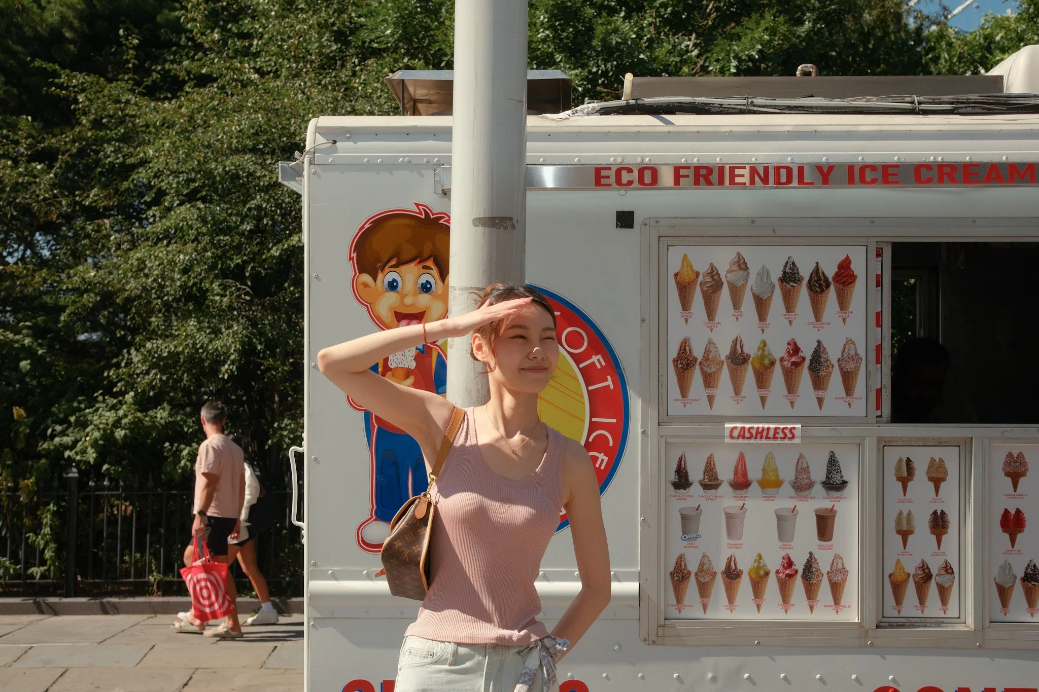 A woman in a pink top salutes in front of an ice cream stand with a large menu of various ice cream cones. There are trees in the background and other people walking nearby.
