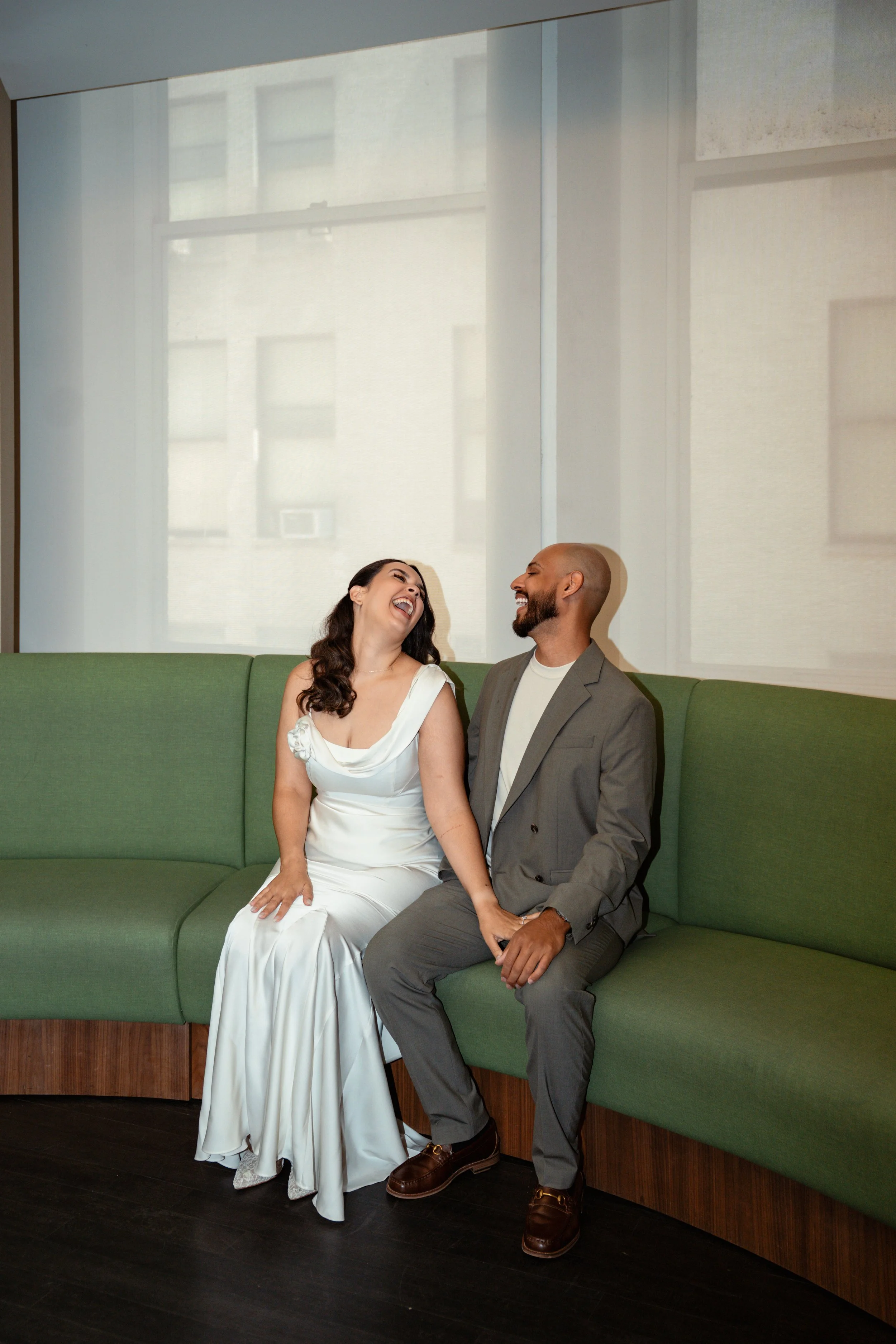 A woman in a white wedding dress and a man in a gray suit are sitting on a green couch, laughing and smiling at each other.