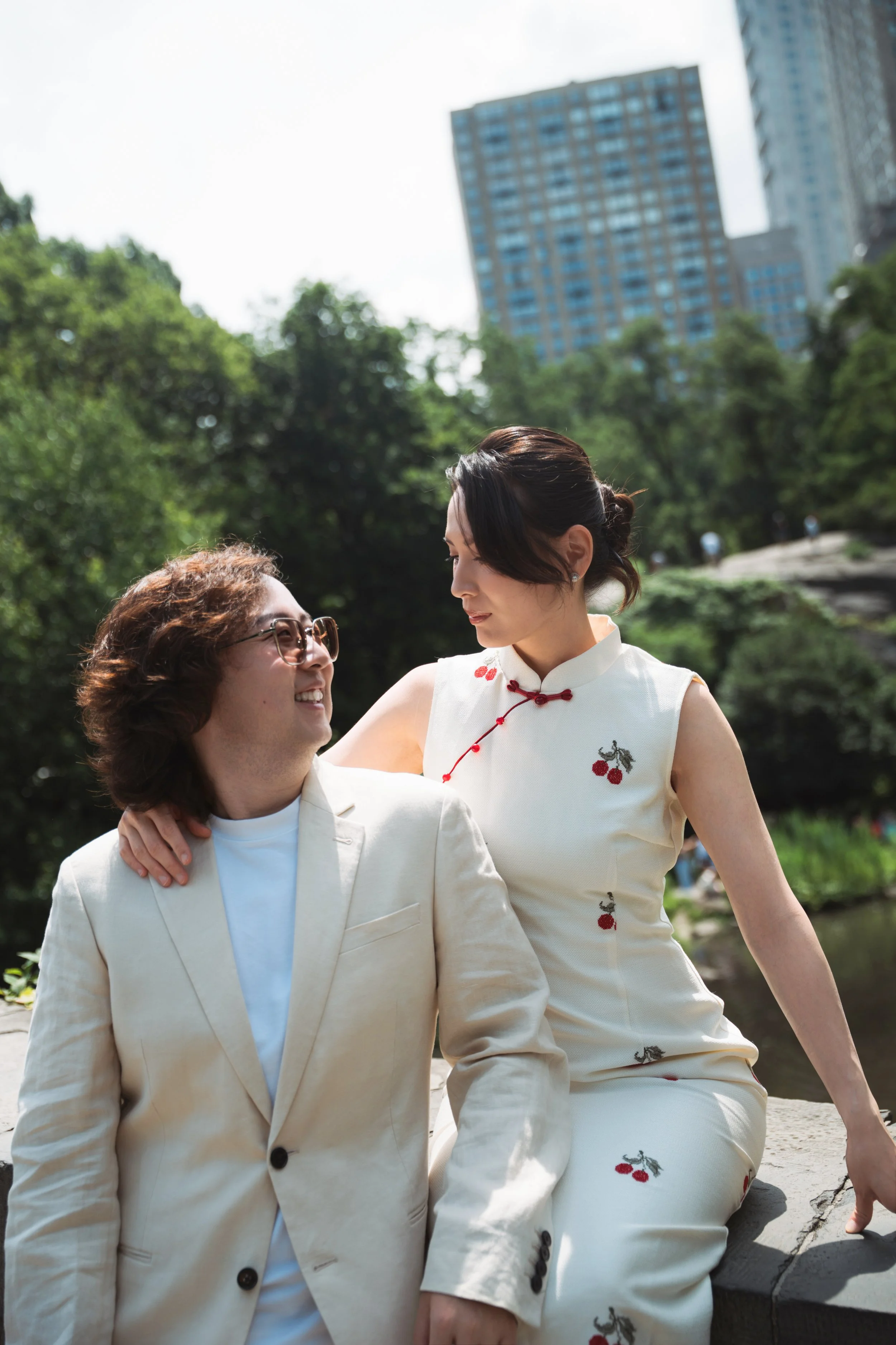 A man with glasses and curly hair and a woman with dark hair in an updo are outdoors, sitting near a river with trees and tall buildings in the background. They are dressed in light-colored clothing with cherry blossom embroidery.