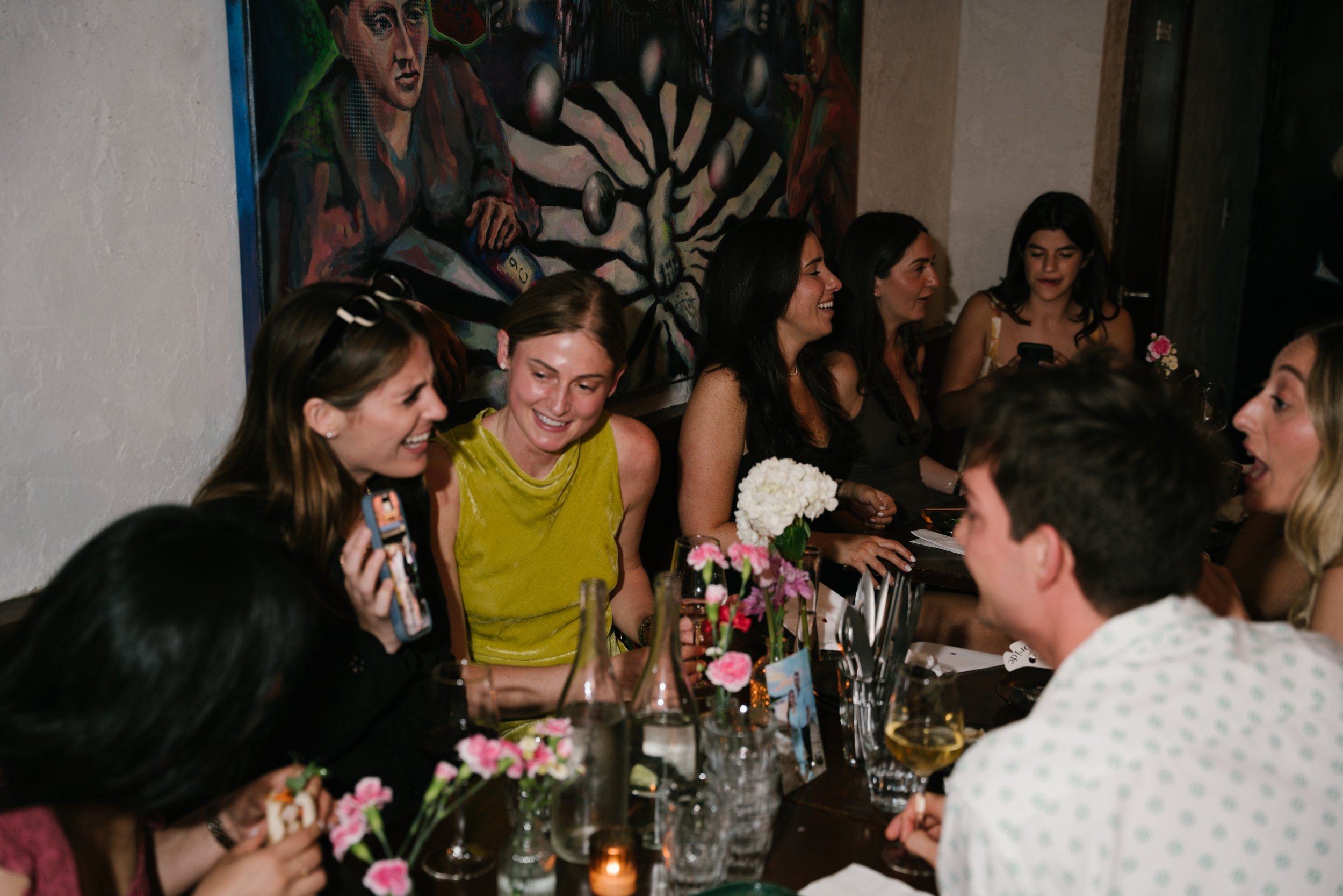 Group of women sitting at a dinner table with flowers and drinks, smiling and talking, with a colorful mural painting in the background.